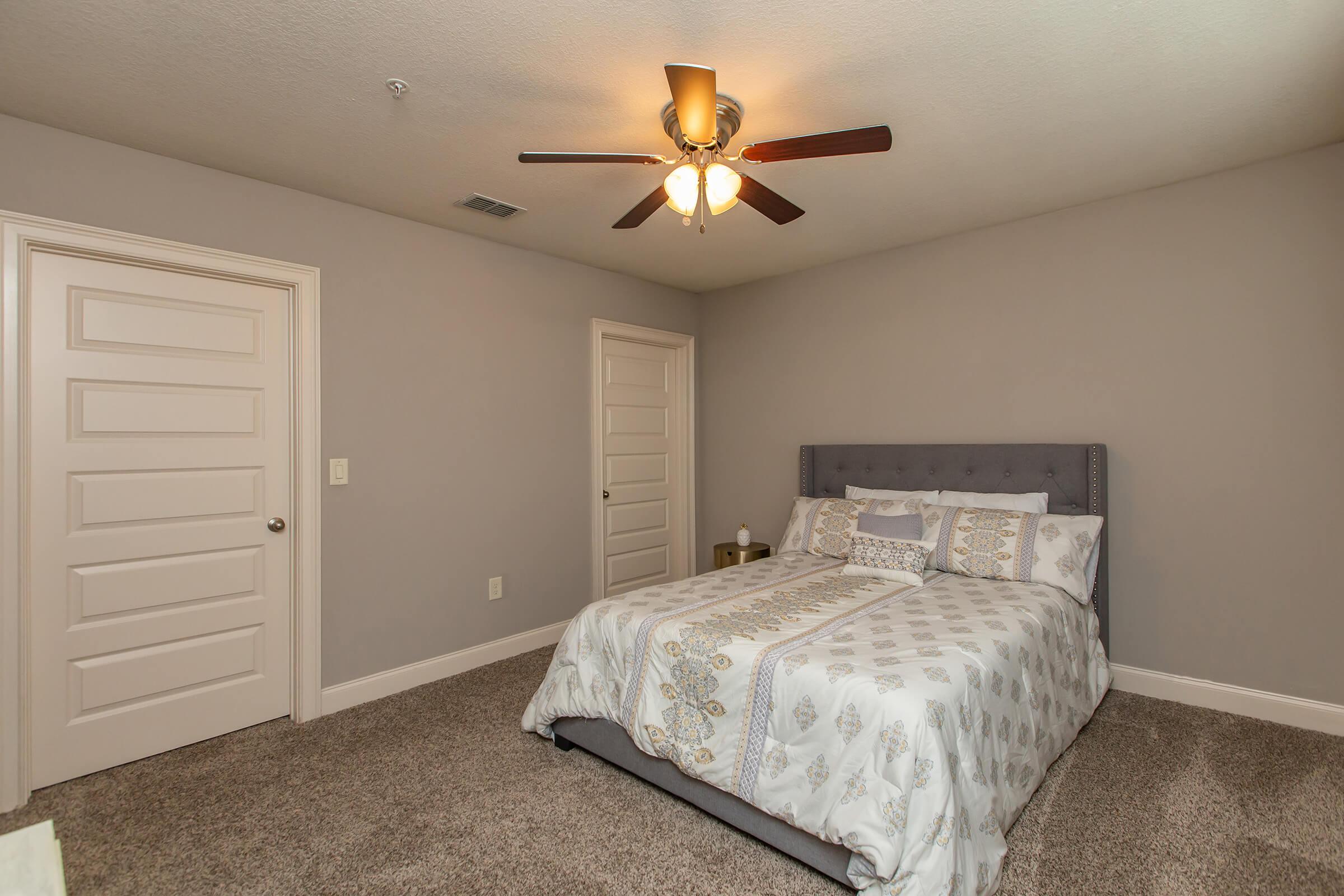 A neatly arranged bedroom featuring a gray upholstered bed with decorative bedding. There is a ceiling fan with light, and two doors leading to other rooms. The walls are painted in a light gray color, and the carpet is a soft beige, creating a cozy atmosphere.