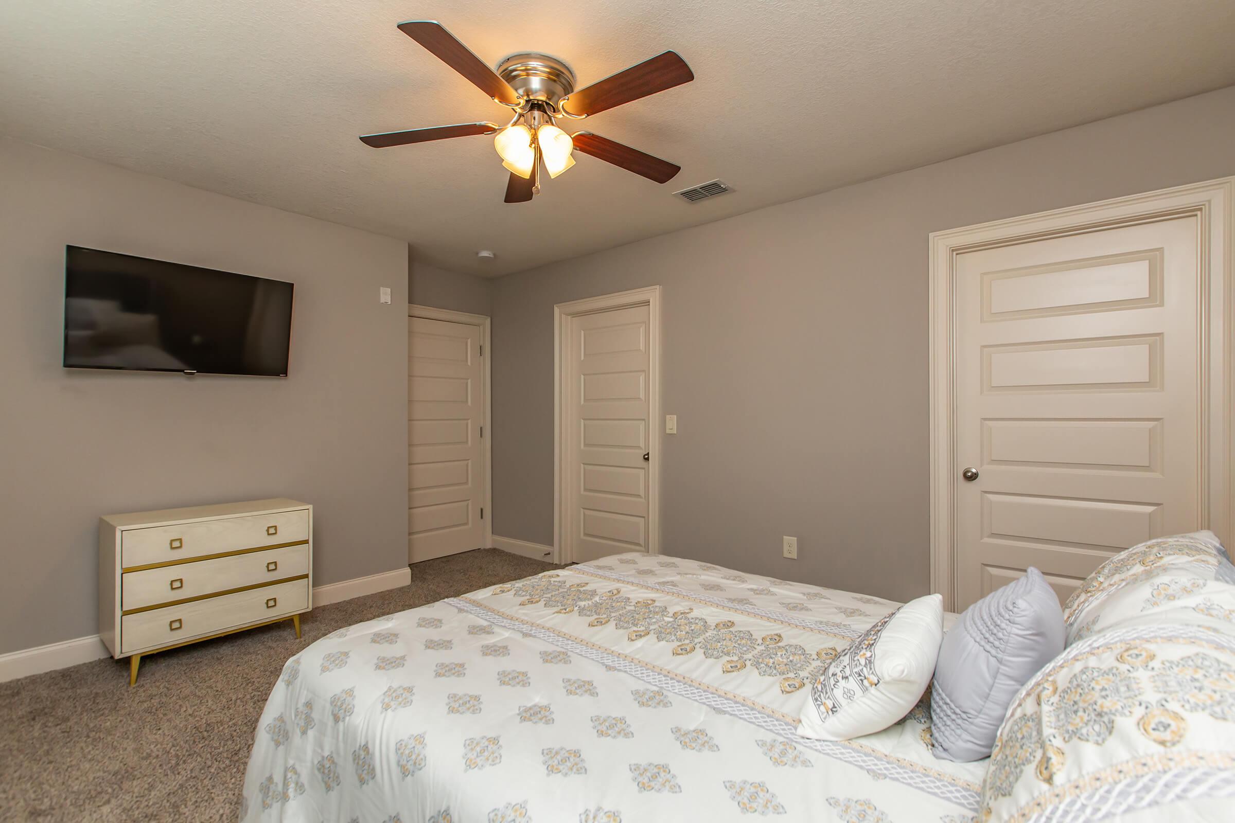 A cozy bedroom featuring a neatly made bed with decorative pillows, a white dresser with gold accents, a flat-screen TV mounted on the wall, and a ceiling fan. The room has light grey walls and plush carpet, with two doors leading to other spaces, creating a serene atmosphere.