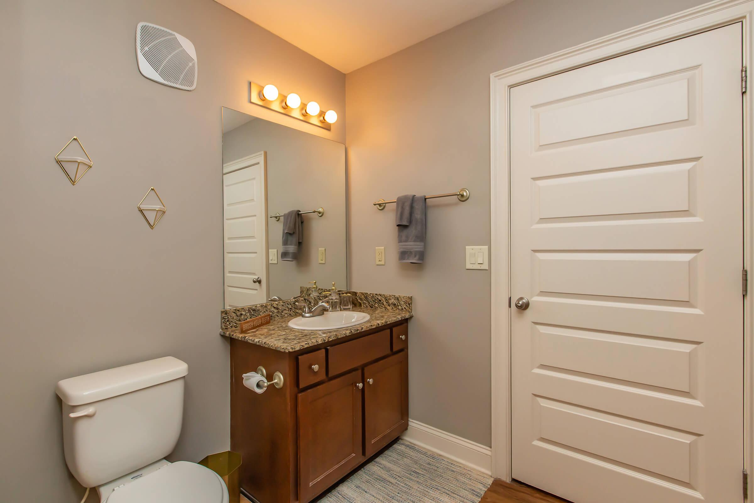 A modern bathroom featuring a light gray wall, a wooden vanity with a granite countertop, a round mirror above the sink, and elegant lighting fixtures. The space includes a white toilet, a towel rack with gray towels, and a door, adding to the clean and contemporary design.