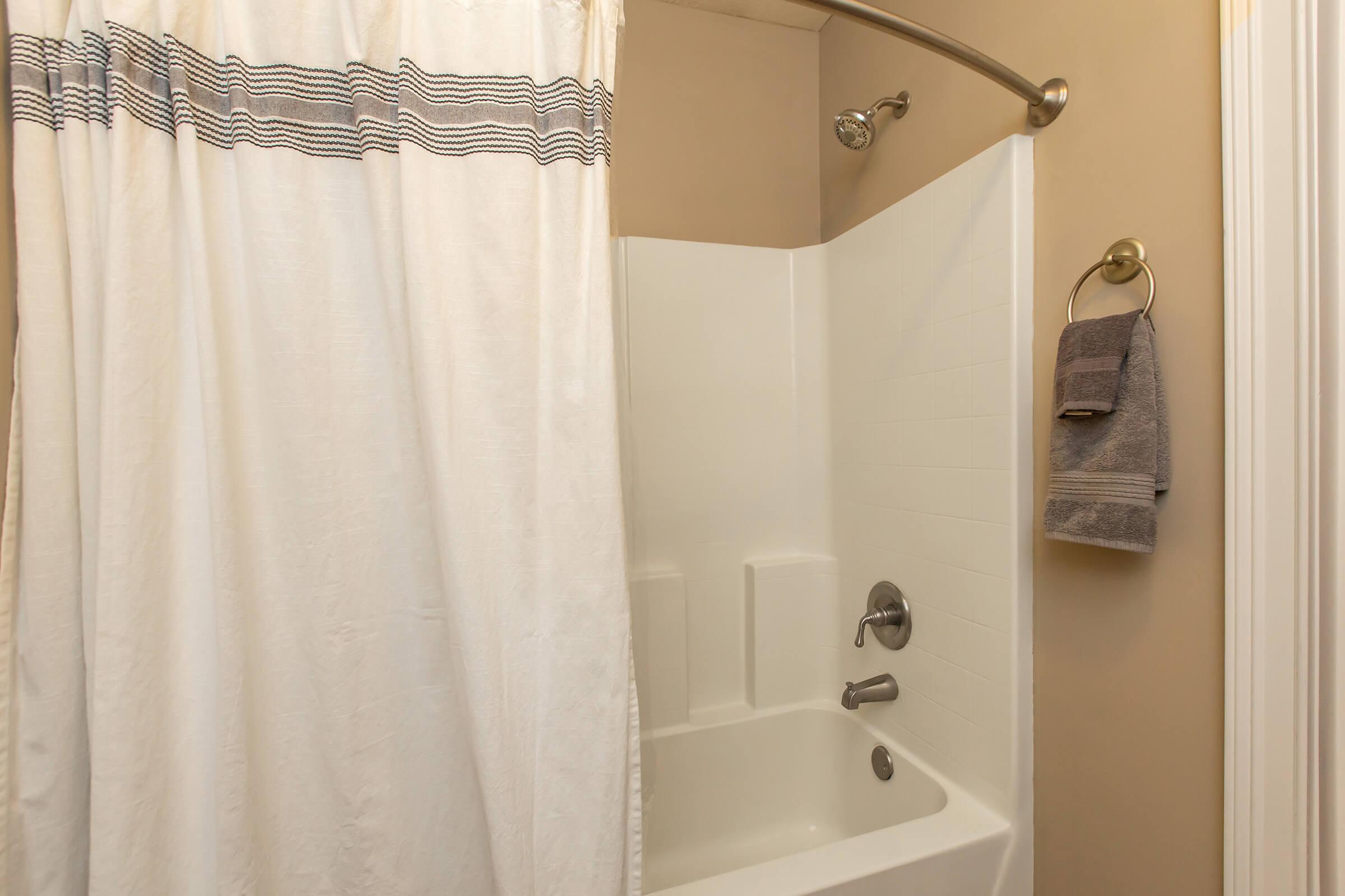 A clean bathroom featuring a white bathtub and shower area. The shower curtain is white with black stripes at the top. A silver towel rack holds a gray towel on the wall nearby. The walls are a light color, enhancing the space's brightness and cleanliness.