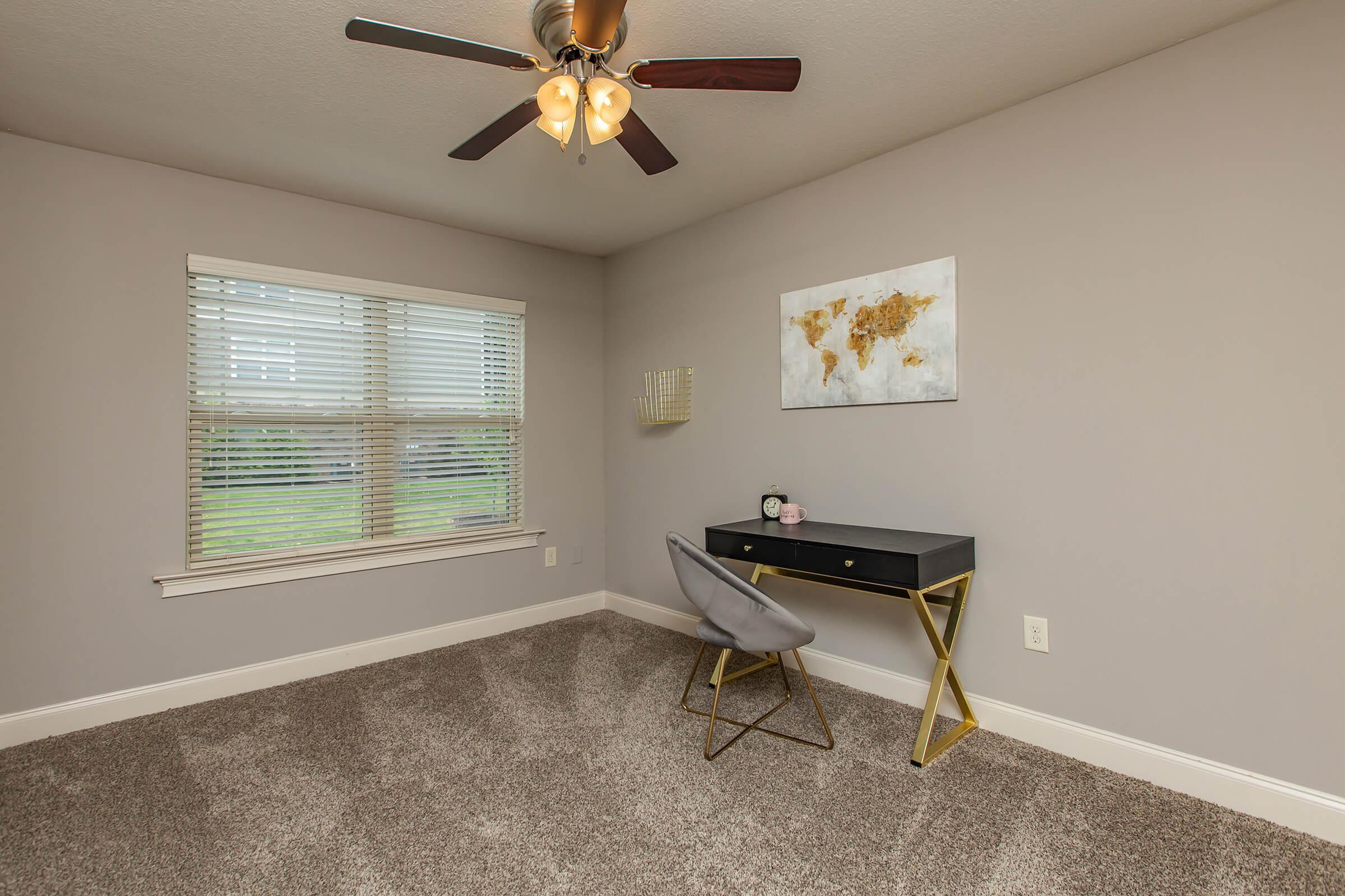 A cozy room featuring a ceiling fan, a large window with blinds, and a stylish desk with a gold-colored frame. The walls are painted in a soft gray, and there is a framed world map hanging on the wall. Plush carpet covers the floor, creating a comfortable atmosphere.