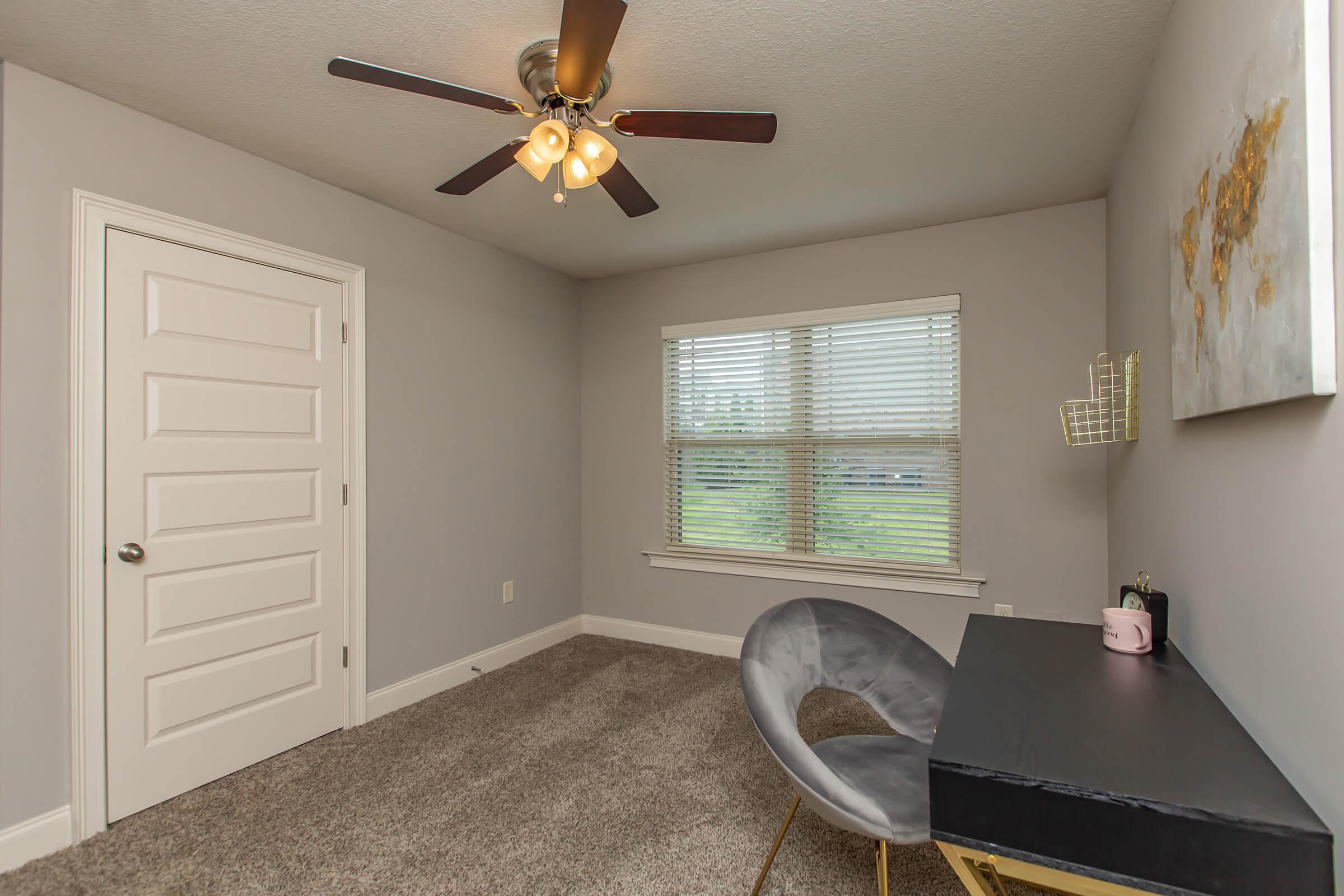 A minimalist room with gray walls, featuring a ceiling fan with light, a window with blinds, a stylish gray chair, and a small black desk. There's a decorative wall piece and a door in the background. The room has a cozy carpeted floor, creating a comfortable and inviting atmosphere.