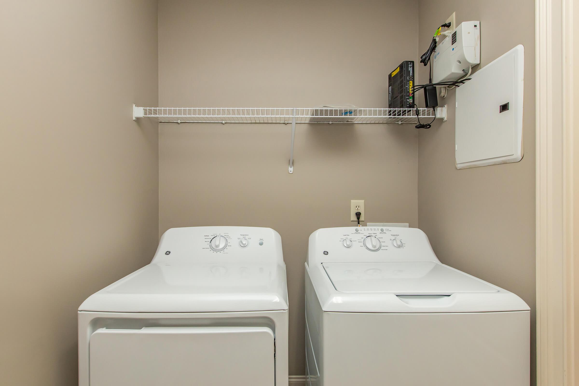 A clean laundry room featuring a white washing machine and a matching dryer side by side. Above them, there is a wire shelf holding a device and a wall-mounted electrical panel. The walls are painted a neutral beige color, creating a simple and functional space.