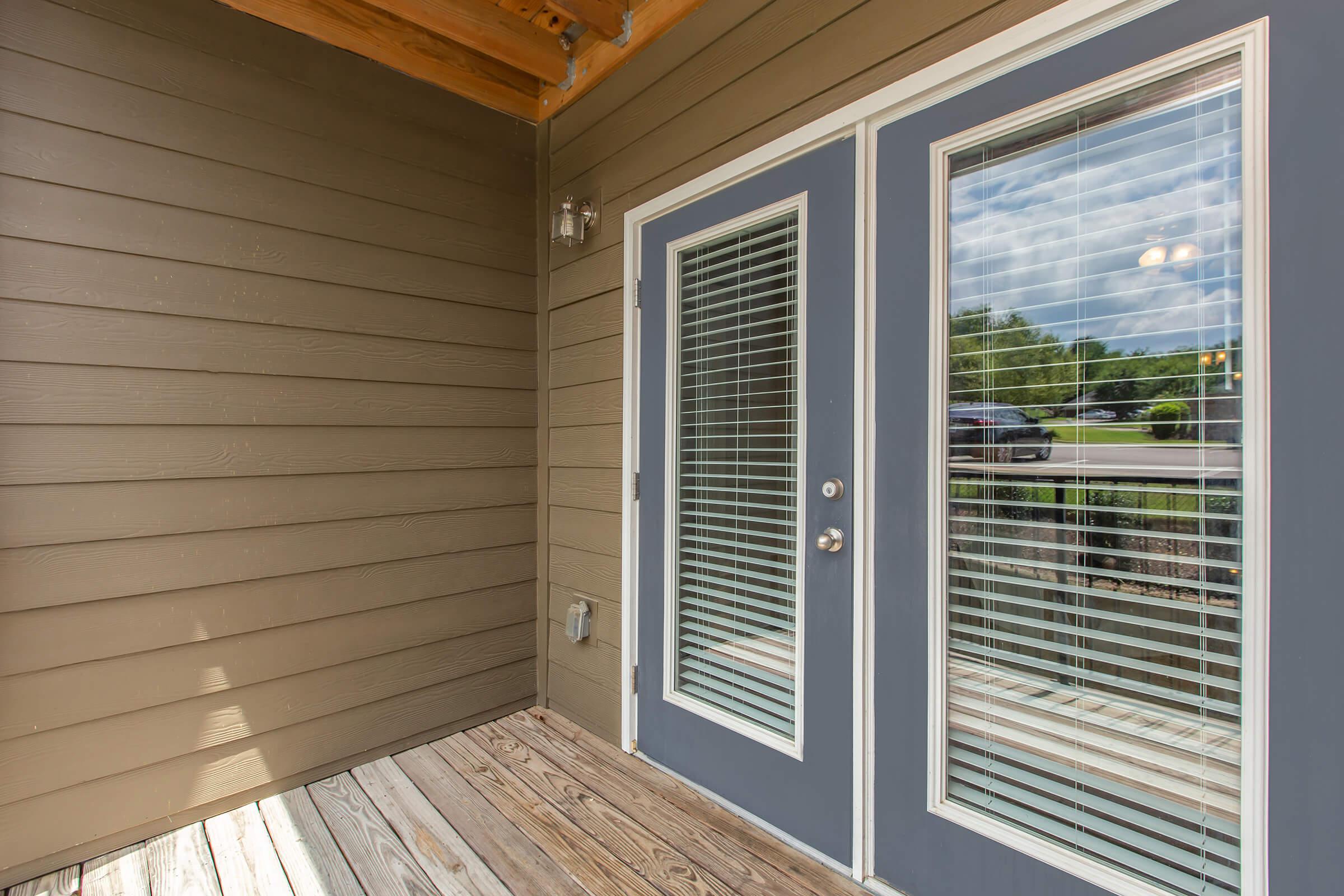 A view of a wooden entryway featuring double doors with vertical blinds, surrounded by a brown exterior wall. The floor is made of wooden boards, and there are large windows beside the doors, allowing natural light to enter. The scene is set in a well-lit, residential area.