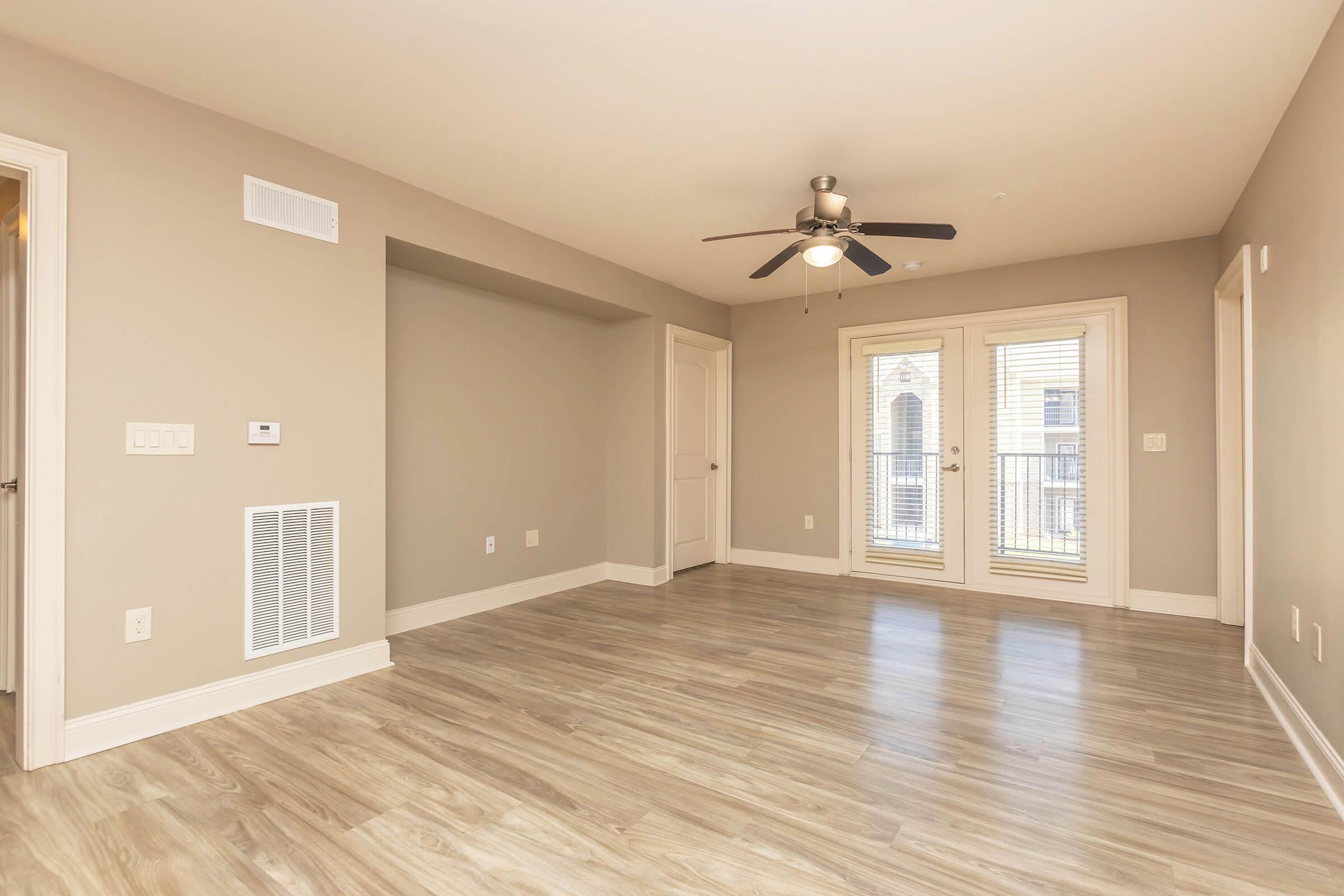 A spacious, unfurnished living room with light gray walls and laminate flooring. It features a ceiling fan, a doorway leading to a balcony with double doors, and a small nook in the wall. The room has natural light from the outdoor access and includes a vent on the wall.