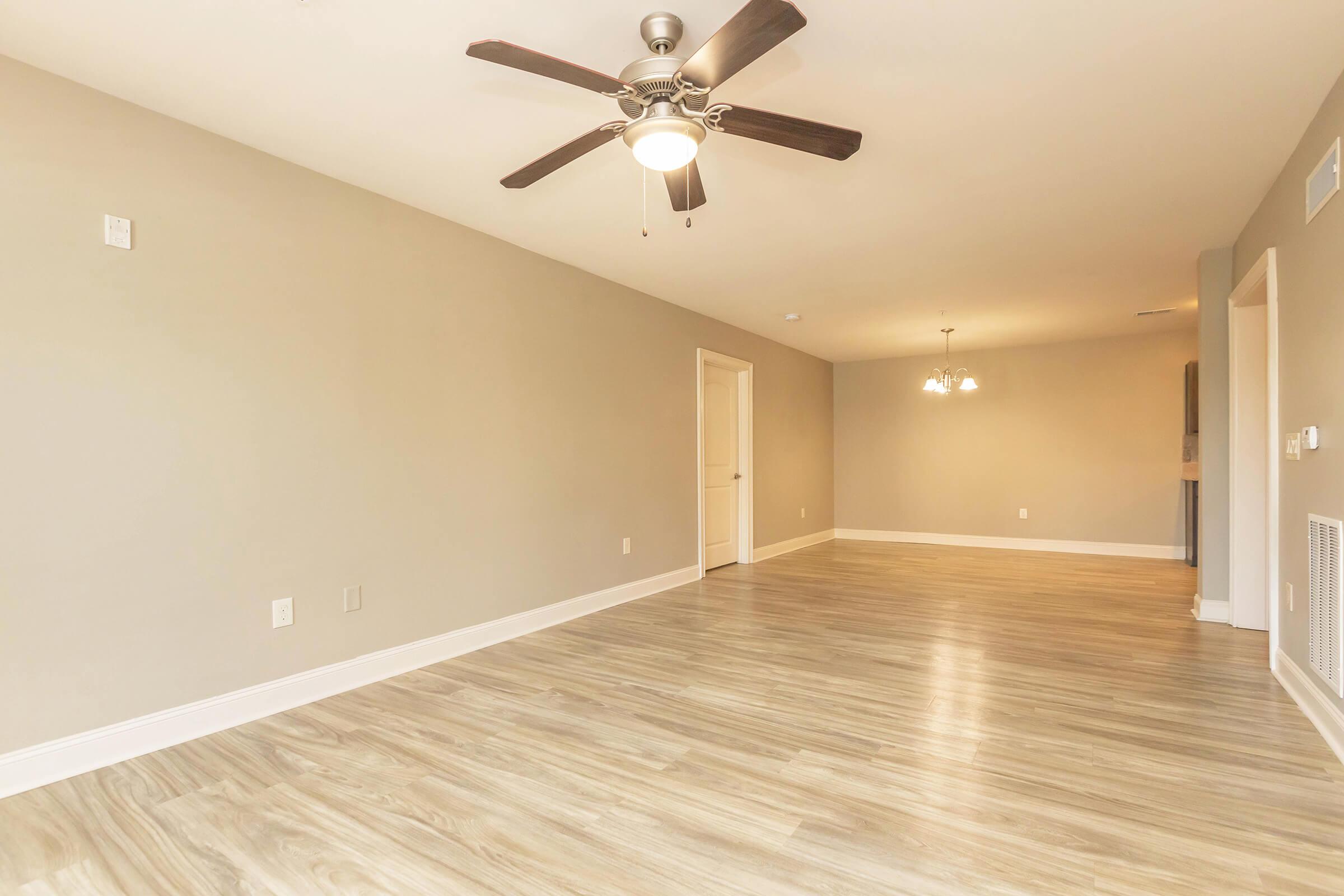 Spacious and empty living room with light gray walls, a ceiling fan with light fixture, and modern wood-look flooring. The room features a doorway leading to another area and a bright chandelier above in a dining space. Natural light brightens the area, creating an inviting atmosphere.