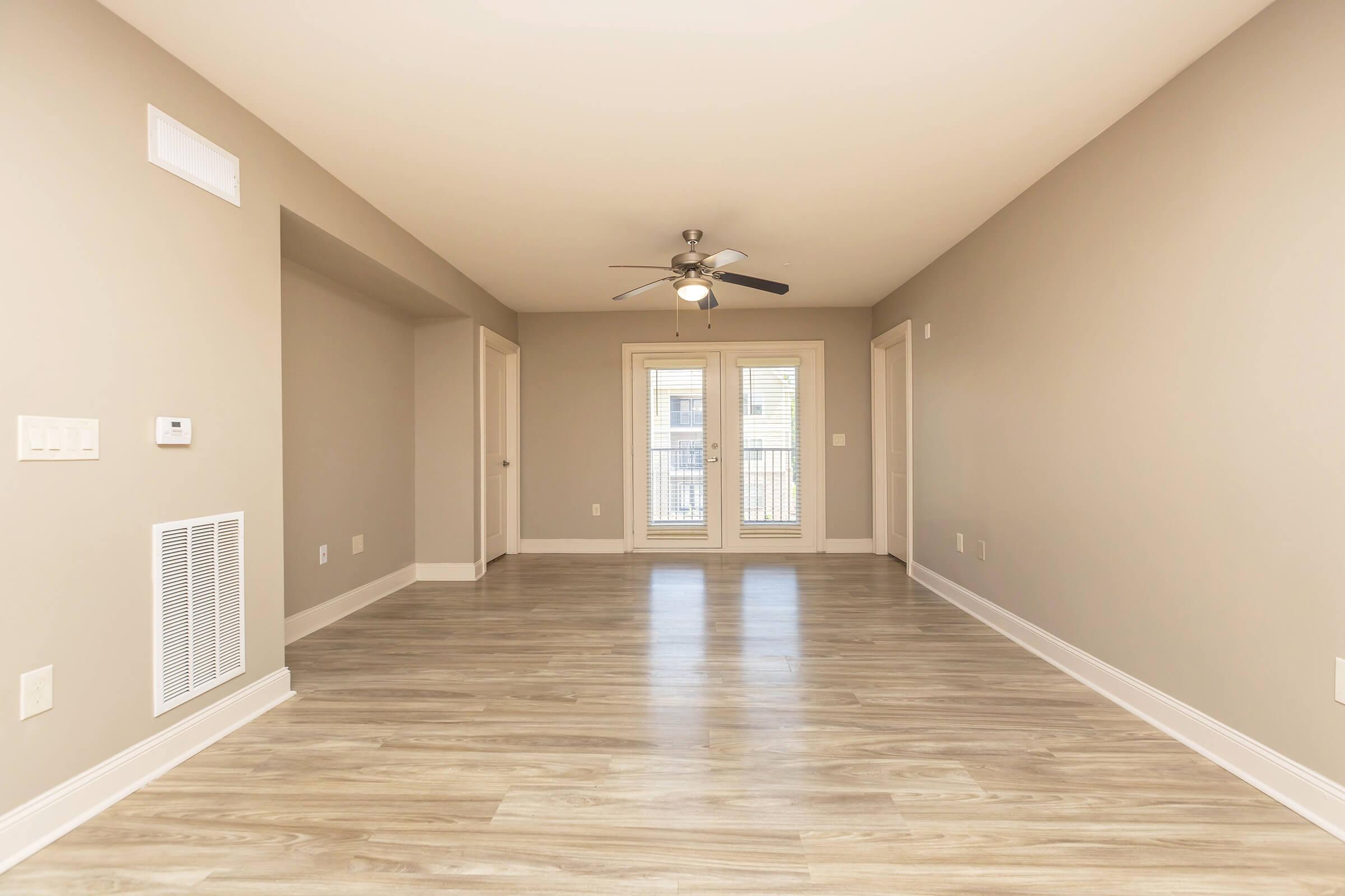 A spacious, empty living room featuring light gray walls and hardwood flooring. A ceiling fan hangs from the ceiling. Large windows with blinds allow natural light to fill the room. Doorways lead to adjacent spaces, and a vent is visible on the wall. The overall atmosphere is bright and open.