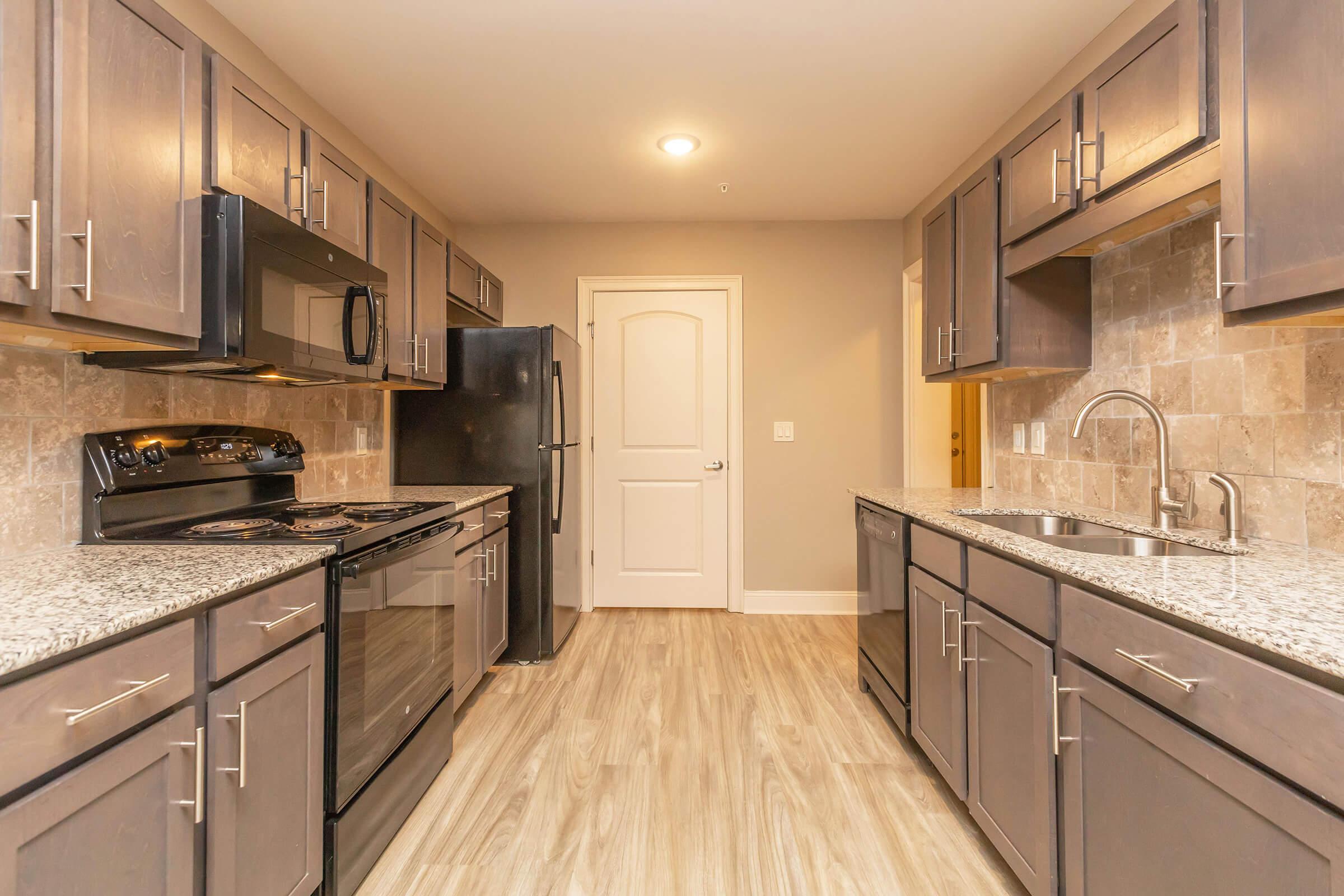 Modern kitchen featuring dark cabinets, stainless steel appliances, and granite countertops. The layout includes a dual sink, a gas stove, an oven, and a refrigerator. Warm lighting enhances the neutral-toned walls and the spacious design creates a welcoming atmosphere. Wood-like flooring completes the contemporary look.