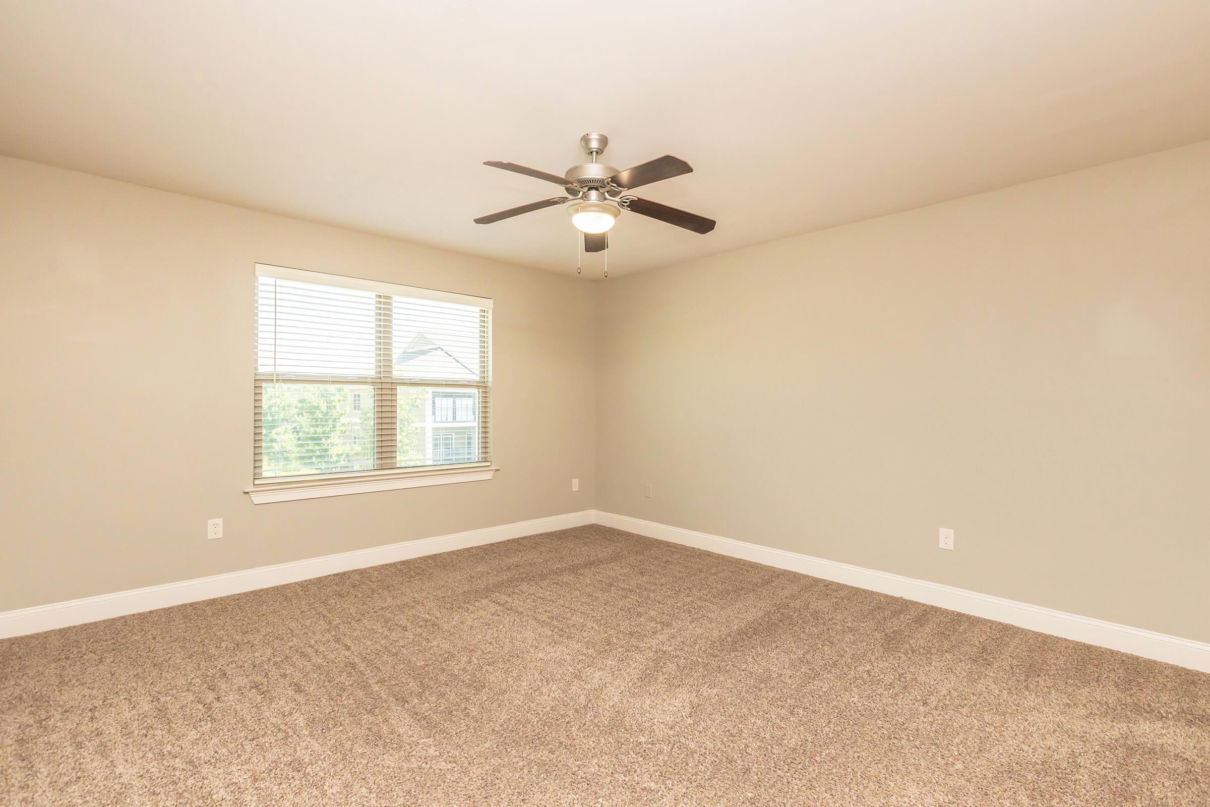 A brightly lit, empty room with beige walls and a plush carpet. A ceiling fan is mounted in the center, and there is a window with blinds on one wall, allowing natural light to enter. The space is clean and unfurnished, providing a blank canvas for decoration.