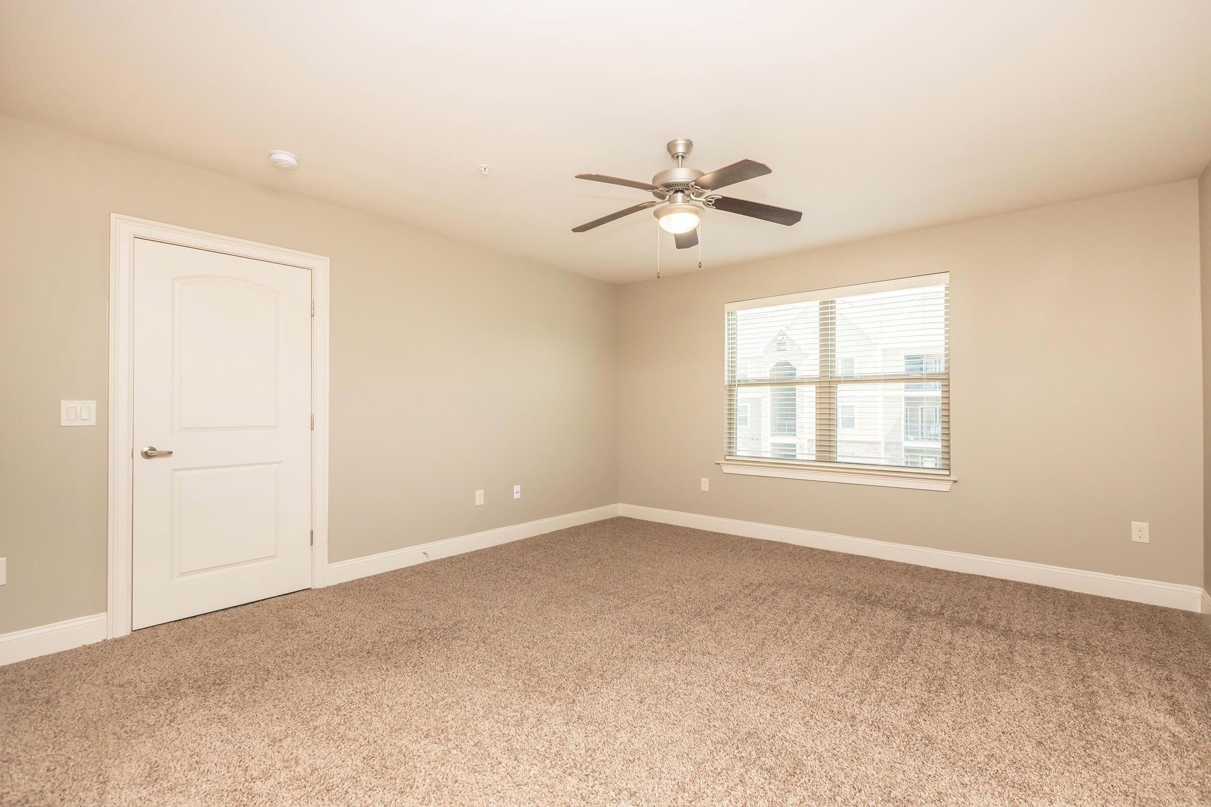 A spacious, empty room with light beige walls and plush carpeting. A ceiling fan is mounted in the center, and a window with blinds allows natural light to enter. The room features a door leading to another space, creating a bright and airy feel.