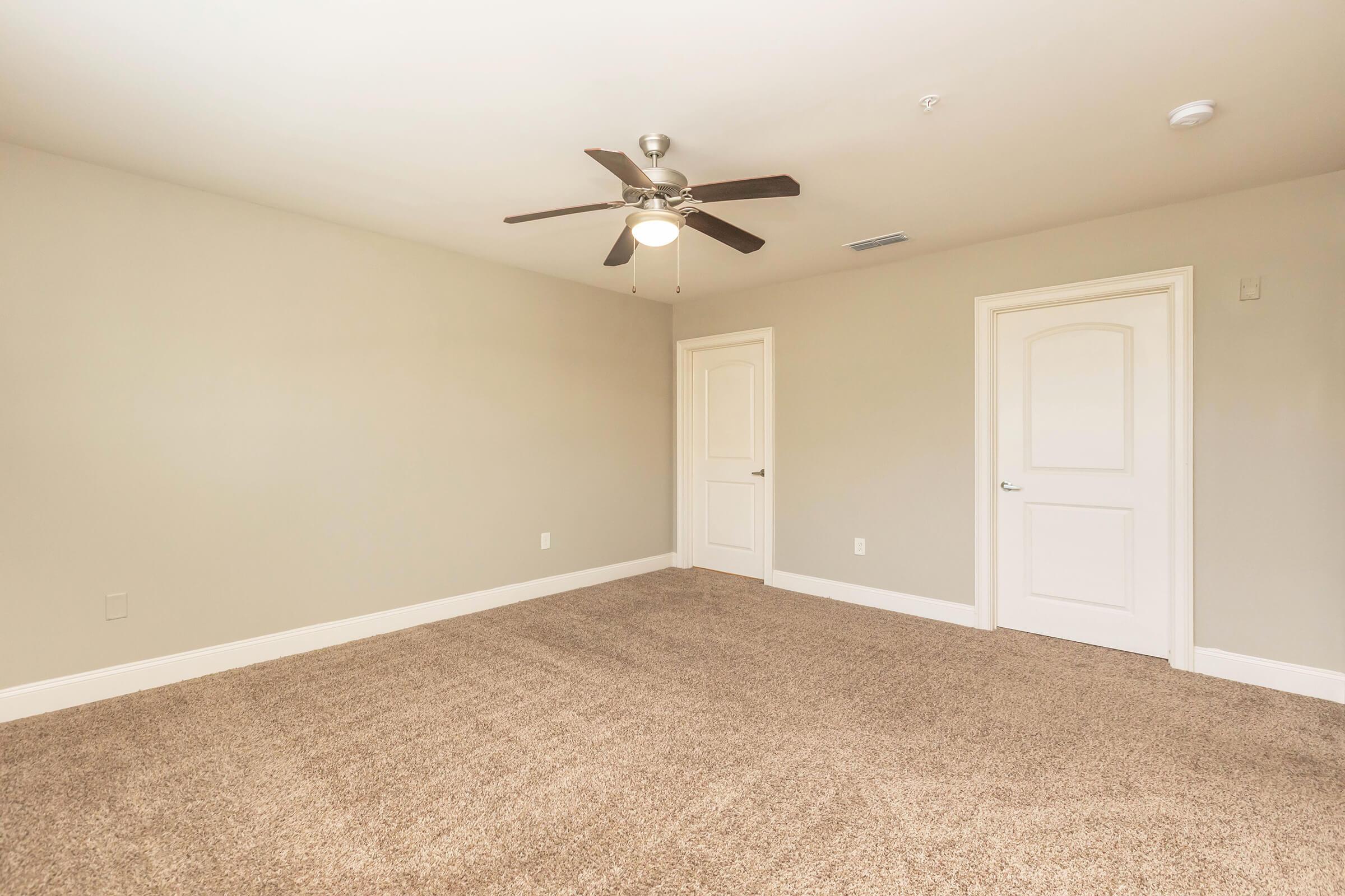 A spacious, empty room with light beige walls and a ceiling fan. The floor is covered in brown carpet, and there are two closed white doors on opposite walls. Natural light filters in, creating a bright and airy atmosphere, ideal for various uses.