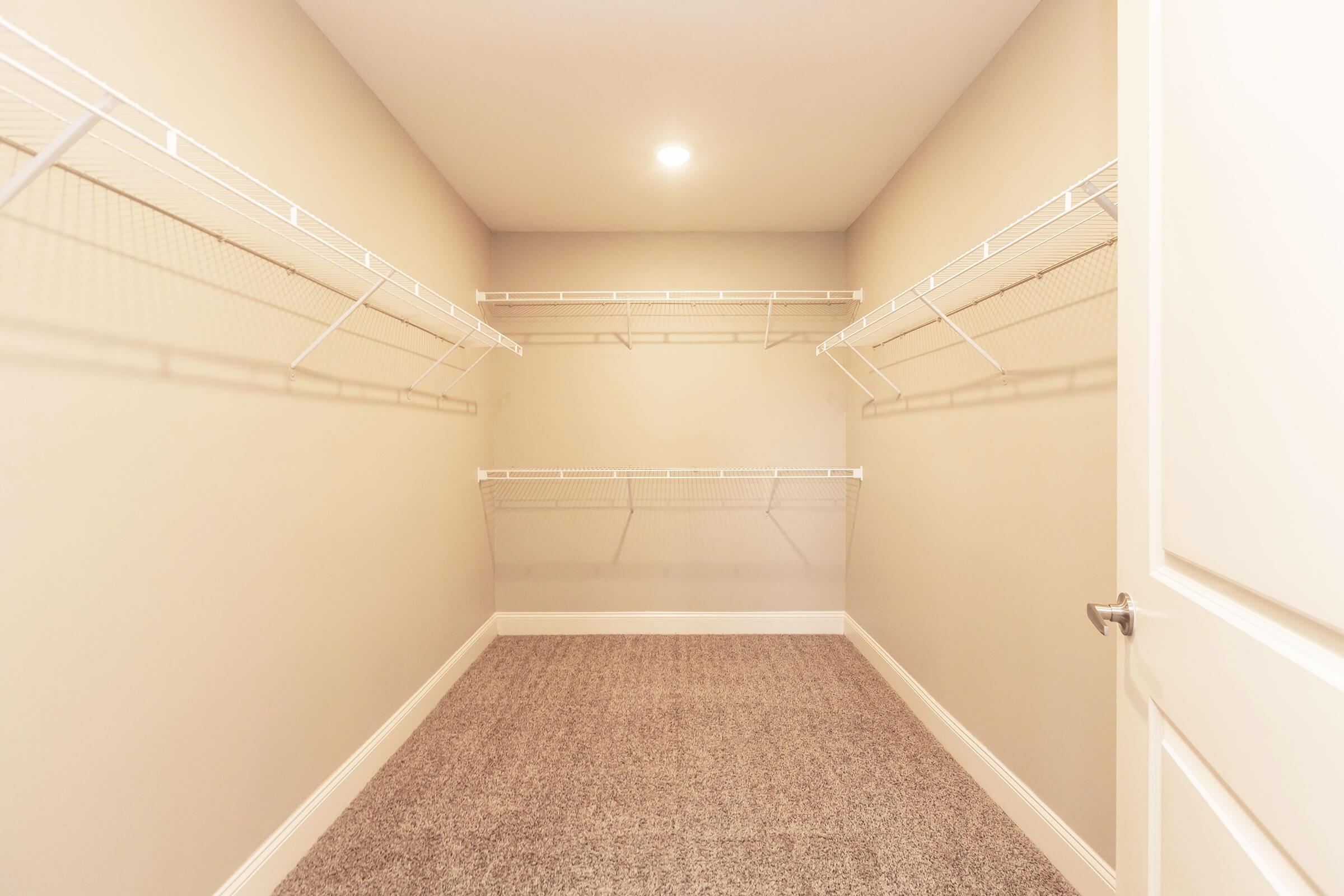 A spacious, empty walk-in closet featuring white wire shelving against light beige walls. The floor is covered with plush carpet, and soft overhead lighting illuminates the area. The closet has a white door slightly ajar, creating a bright and open atmosphere.