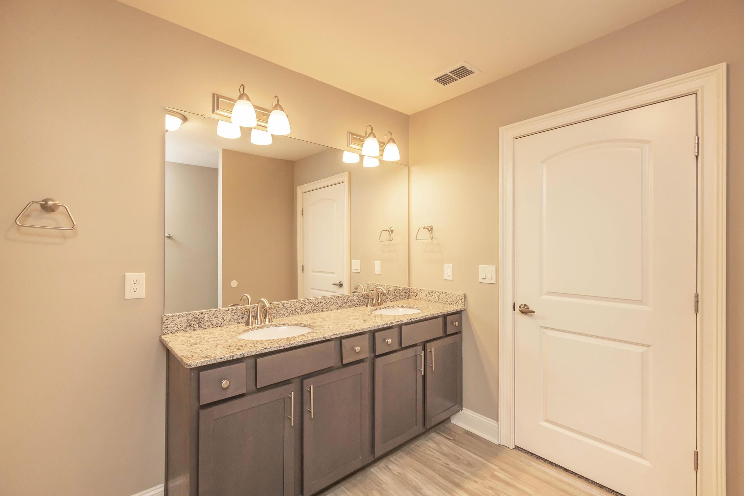 A modern bathroom featuring a double vanity with a granite countertop, two sinks, wall-mounted mirrors, and pendant lighting. The walls are painted in a neutral tone, and there's a stylish door leading to an adjacent space. A towel rack is mounted next to the sinks. The flooring is light-colored laminate.