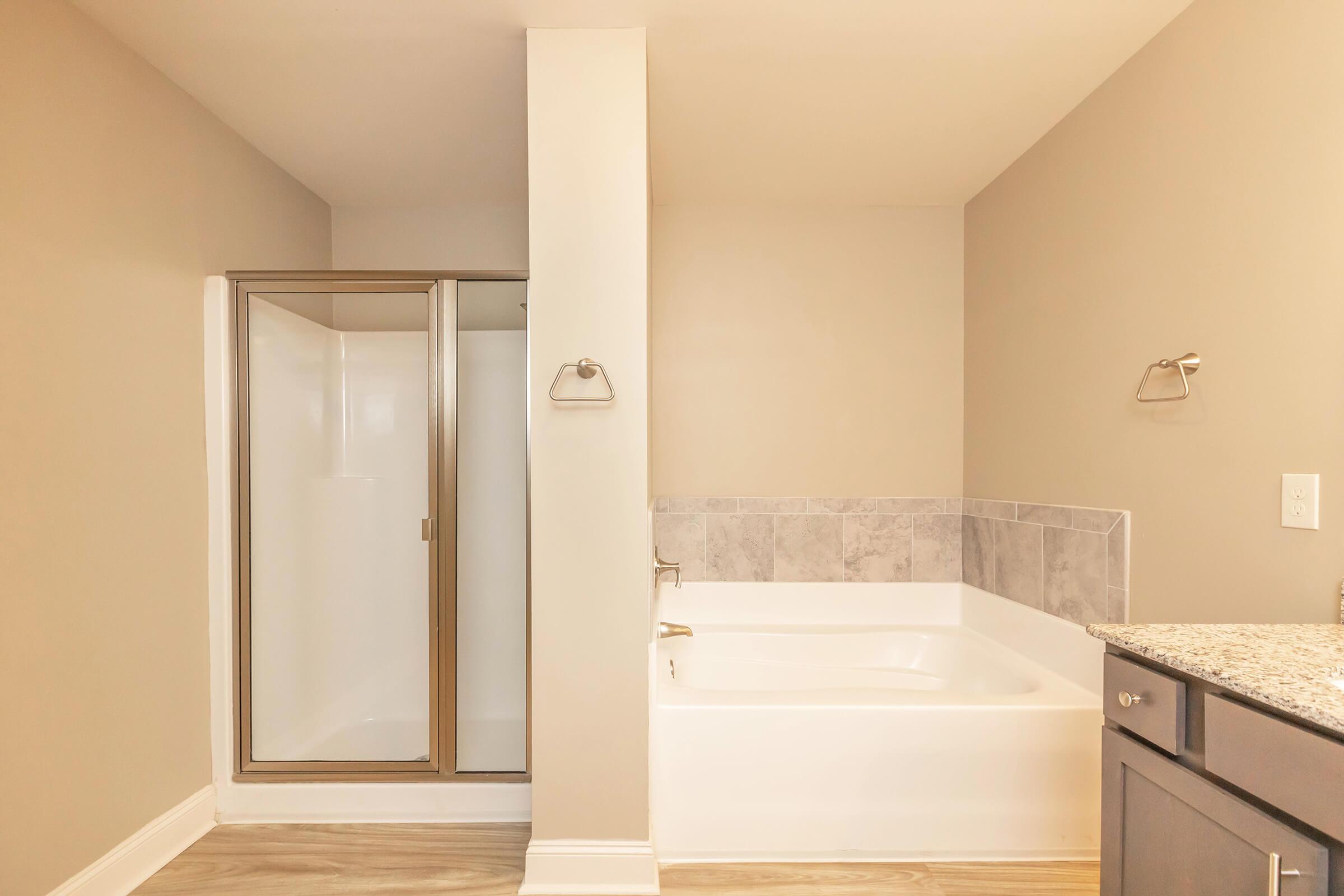 A modern bathroom featuring a glass shower enclosure on the left and a white soaking tub on the right. The walls are painted soft beige, and the flooring is a light wood look. A granite countertop is visible beneath a sink, with silver towel bars fixed to the wall for added convenience.