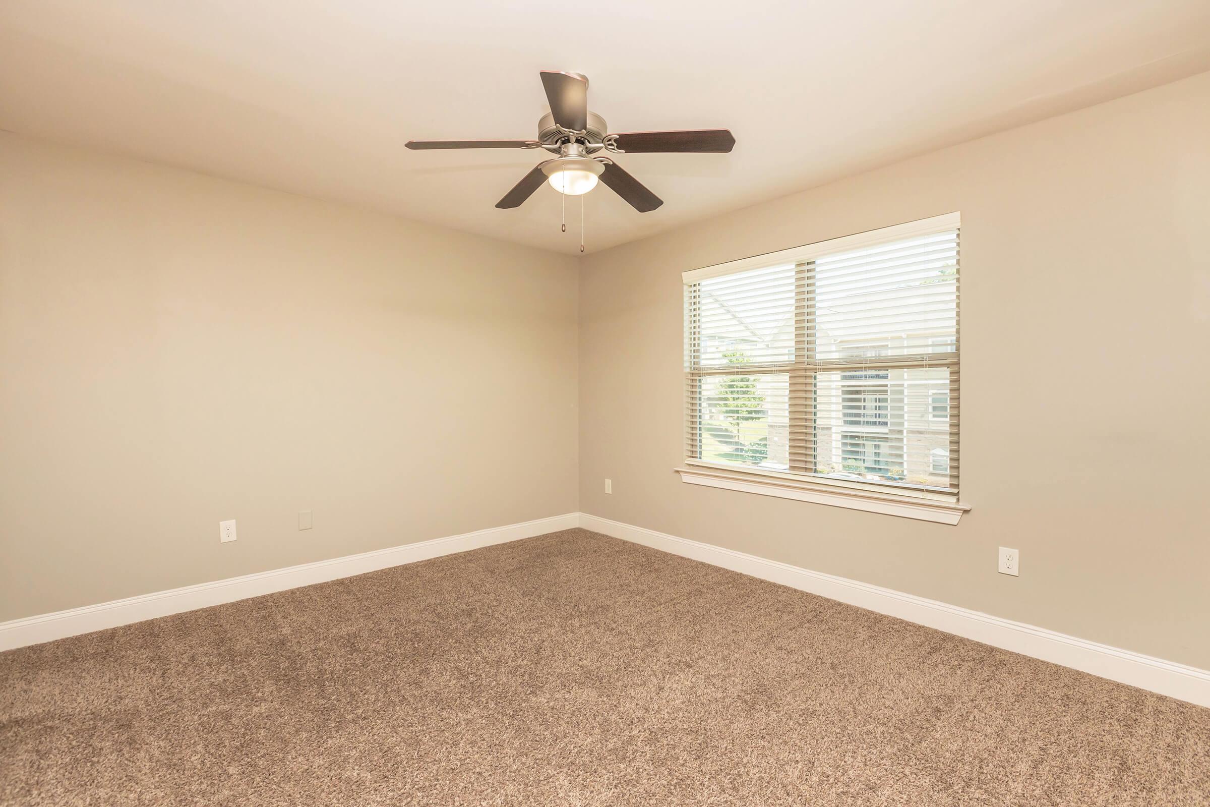 A spacious, empty room with light beige walls and a ceiling fan. Natural light enters through a large window with white blinds, offering a view of an outdoor area. The floor is covered in plush, light brown carpet, creating a warm atmosphere.