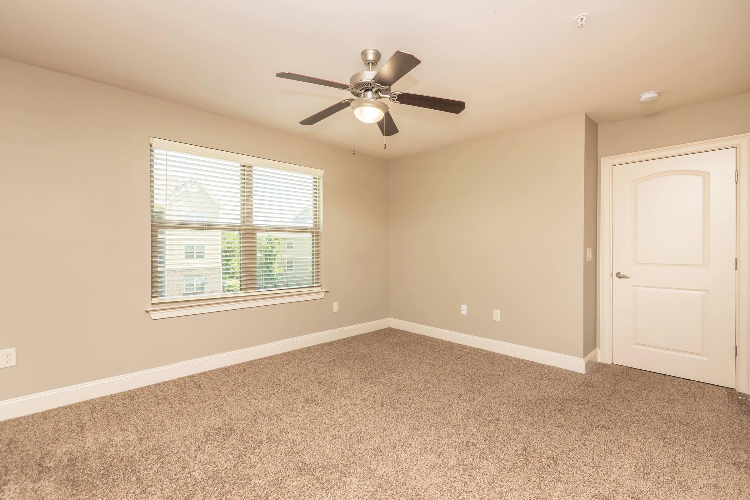 A spacious, empty bedroom featuring beige walls and carpet. A ceiling fan is installed, and natural light streams in through a window with blinds. There is a light-colored door on the right side, completing the simple and clean aesthetic of the room.