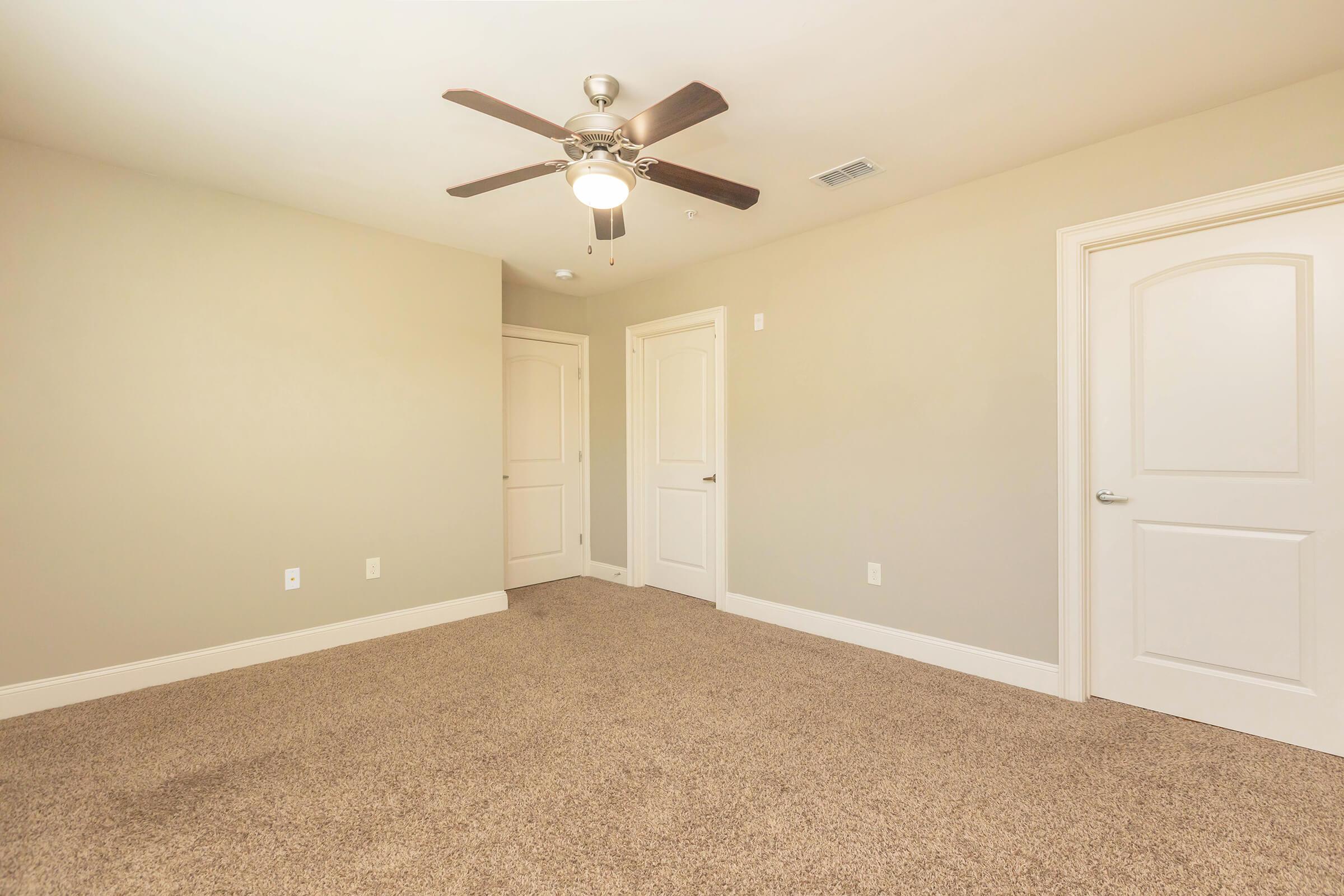 Empty room with light beige walls and carpeted floor. A ceiling fan is mounted in the center, and two doors are visible on the right side, one partially open. Natural light brightens the space, creating a neutral and inviting atmosphere.