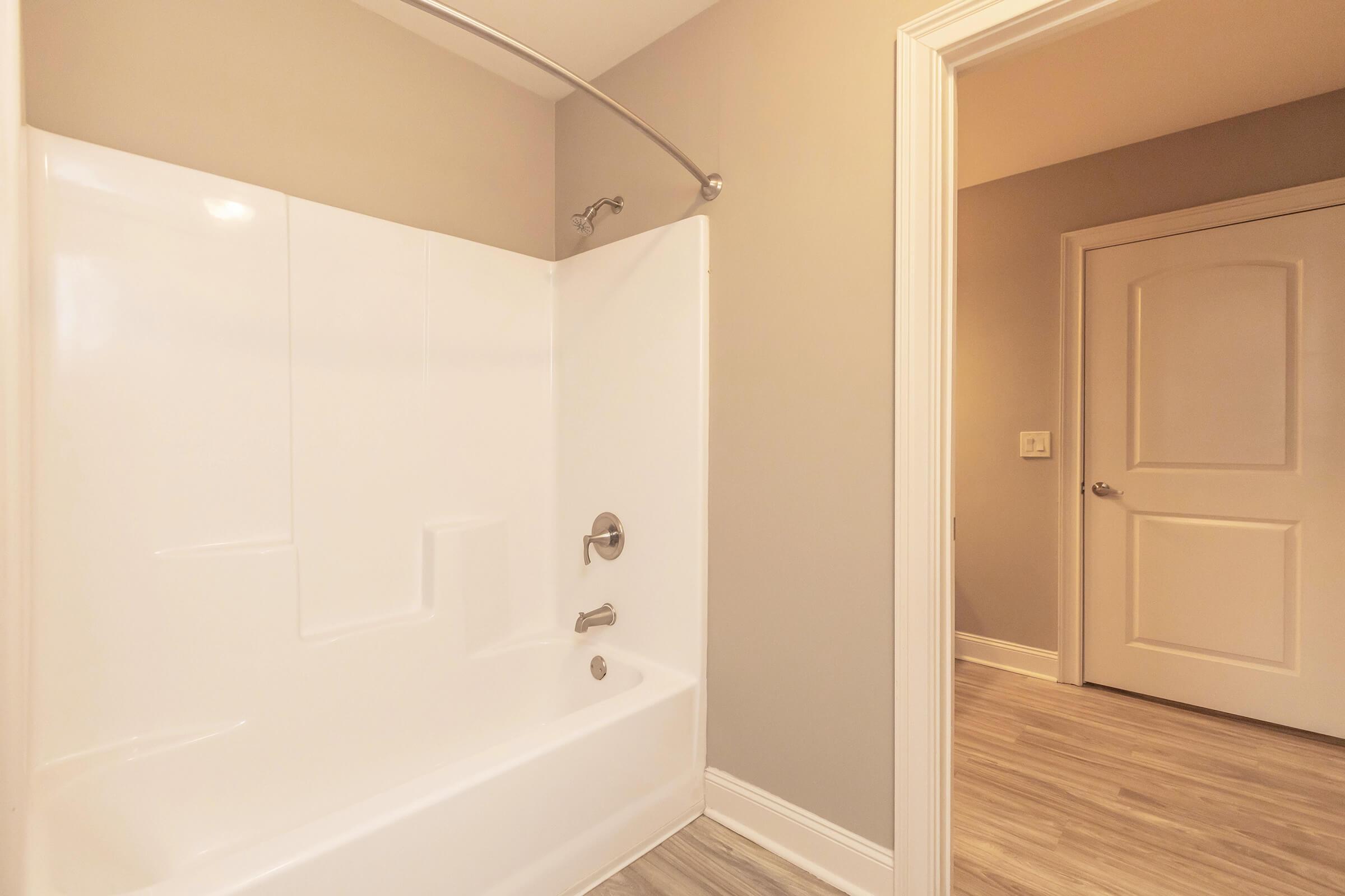 A clean and modern bathroom featuring a white bathtub with a showerhead, light gray walls, and a door leading to another room. The flooring is a light wood laminate, adding warmth to the space. The overall ambiance is bright and inviting.