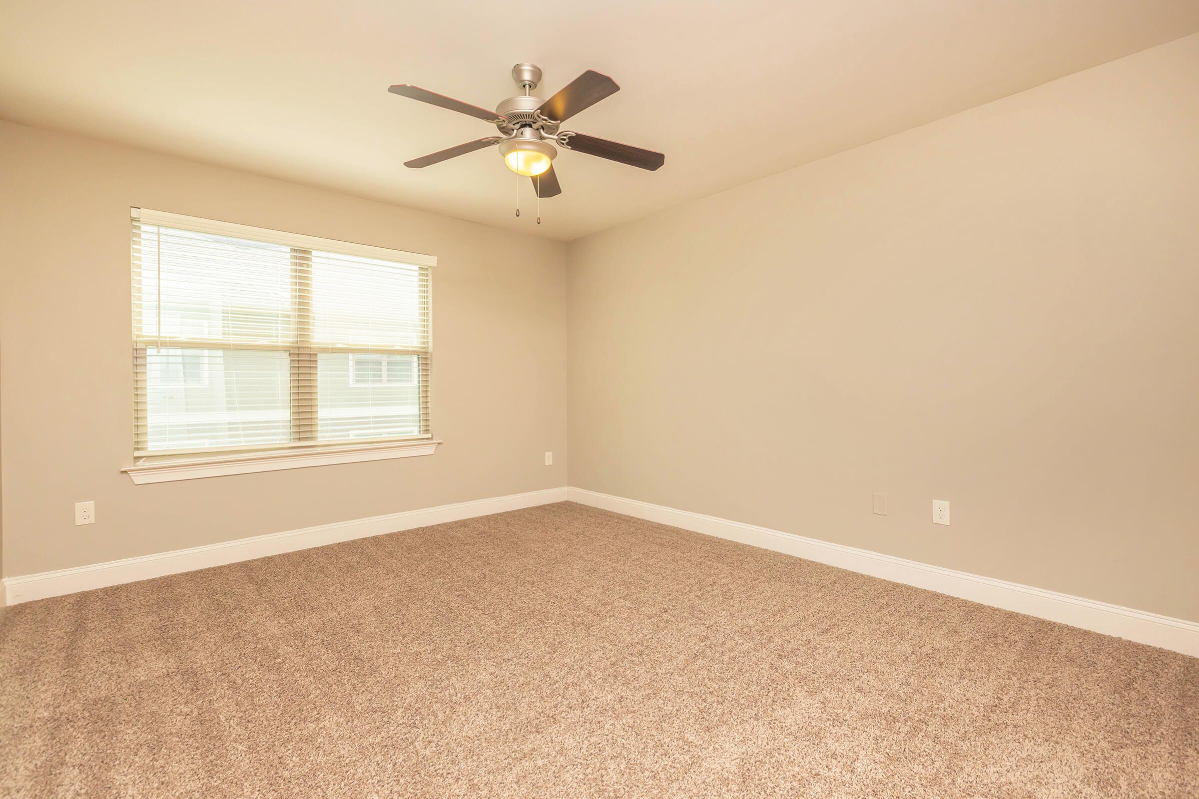 A spacious, empty room featuring light beige walls, a ceiling fan with a light fixture, and a large window with blinds. The floor is covered in plush, tan carpeting, providing a cozy atmosphere. Natural light fills the room, enhancing its openness and potential for various layouts.