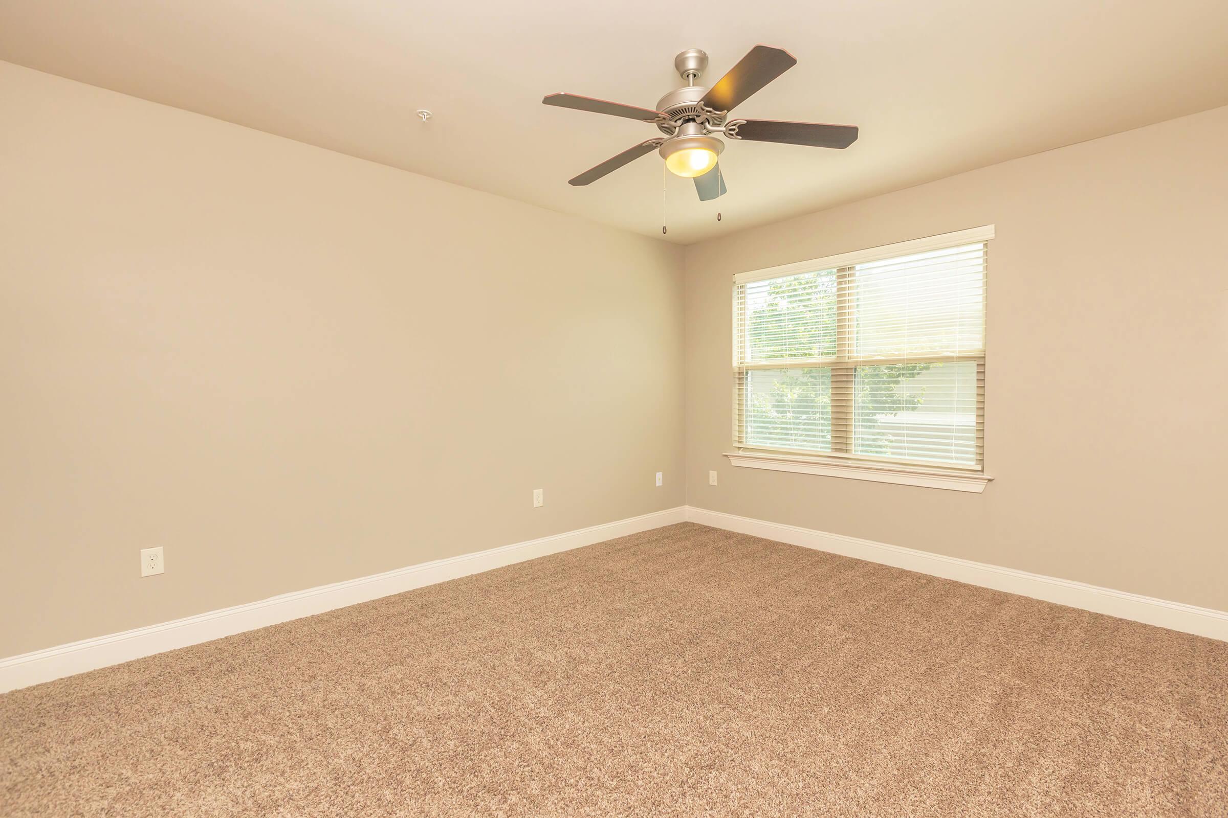 A spacious empty room with beige walls and light brown carpet. It features a ceiling fan with a light fixture, and a large window with white blinds that allows natural light to enter. The room has minimal furnishings and a clean, uncluttered look.