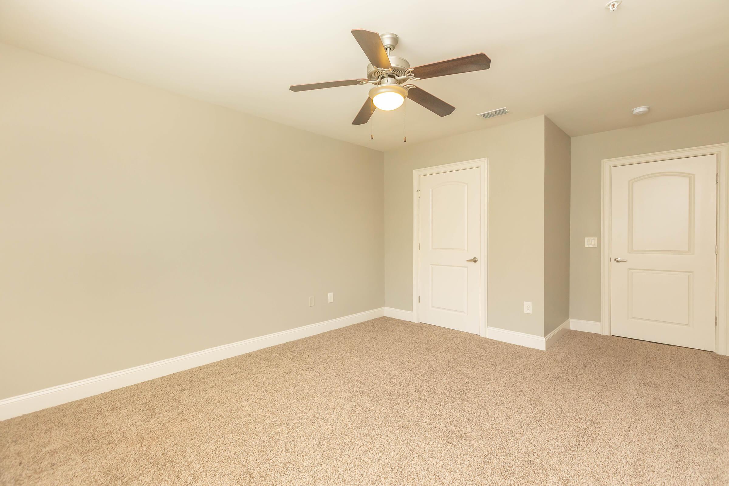 Empty room featuring beige walls and a light-colored carpet. A ceiling fan is mounted in the center, and there are two doors visible: one leading to another area and the other possibly a closet. The overall atmosphere is neutral and spacious, with ample natural light.