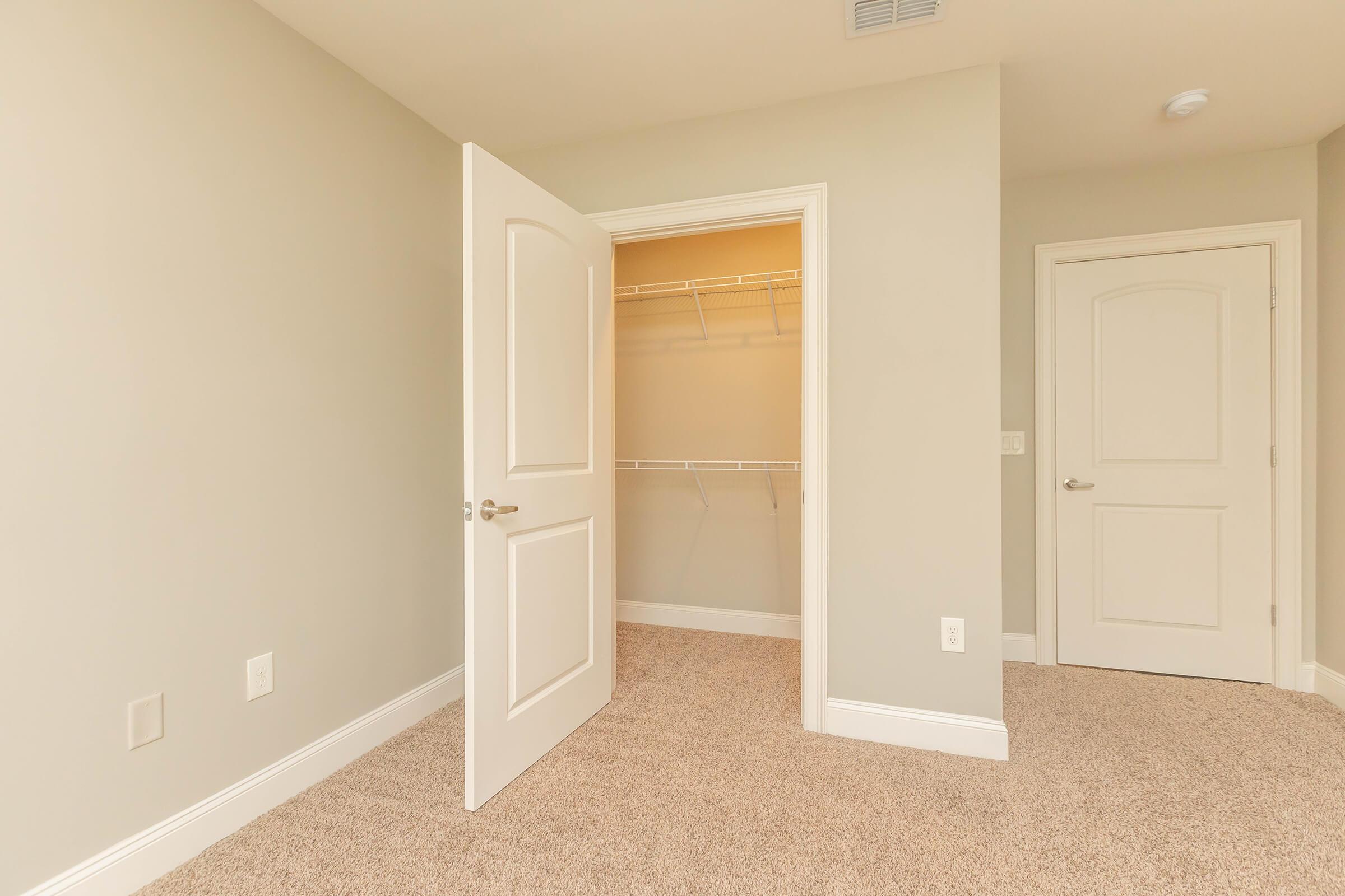 Interior view of a room featuring two doors. One door is open, revealing an empty closet with wire shelving. The walls are painted light gray, and the floor is covered in beige carpeting. The other door is closed, and the overall lighting is bright and inviting.