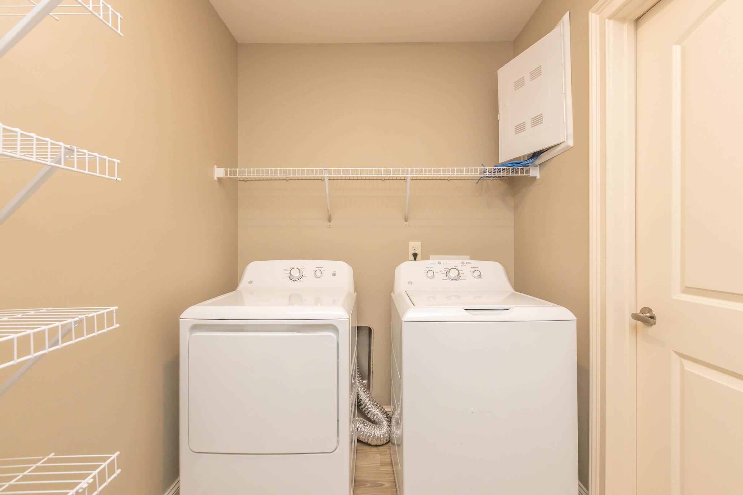 A compact laundry room featuring a white washing machine and dryer side by side against a light beige wall. Above them is a wire shelf for storage, and there is a door leading to another room. The floor is made of light wood-style tiles.