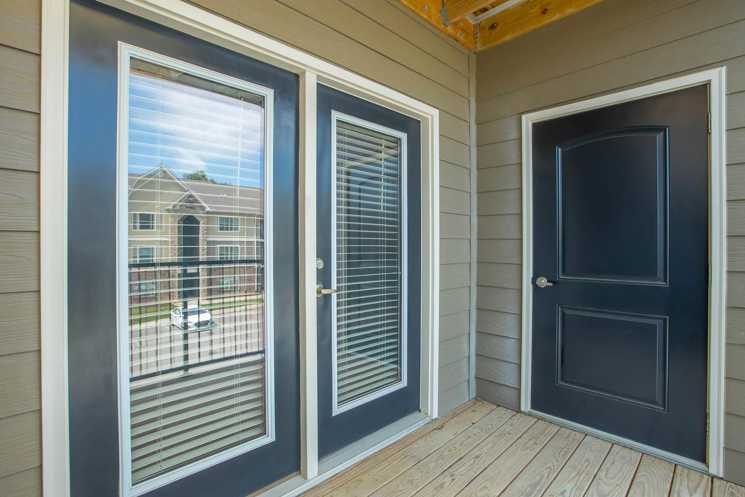 A view of a porch featuring two sets of glass door panels with blinds, adjacent to a solid dark door. The exterior walls are light gray with horizontal siding, and the floor is wooden. In the background, a residential building is visible through the glass doors, suggesting a welcoming entrance.