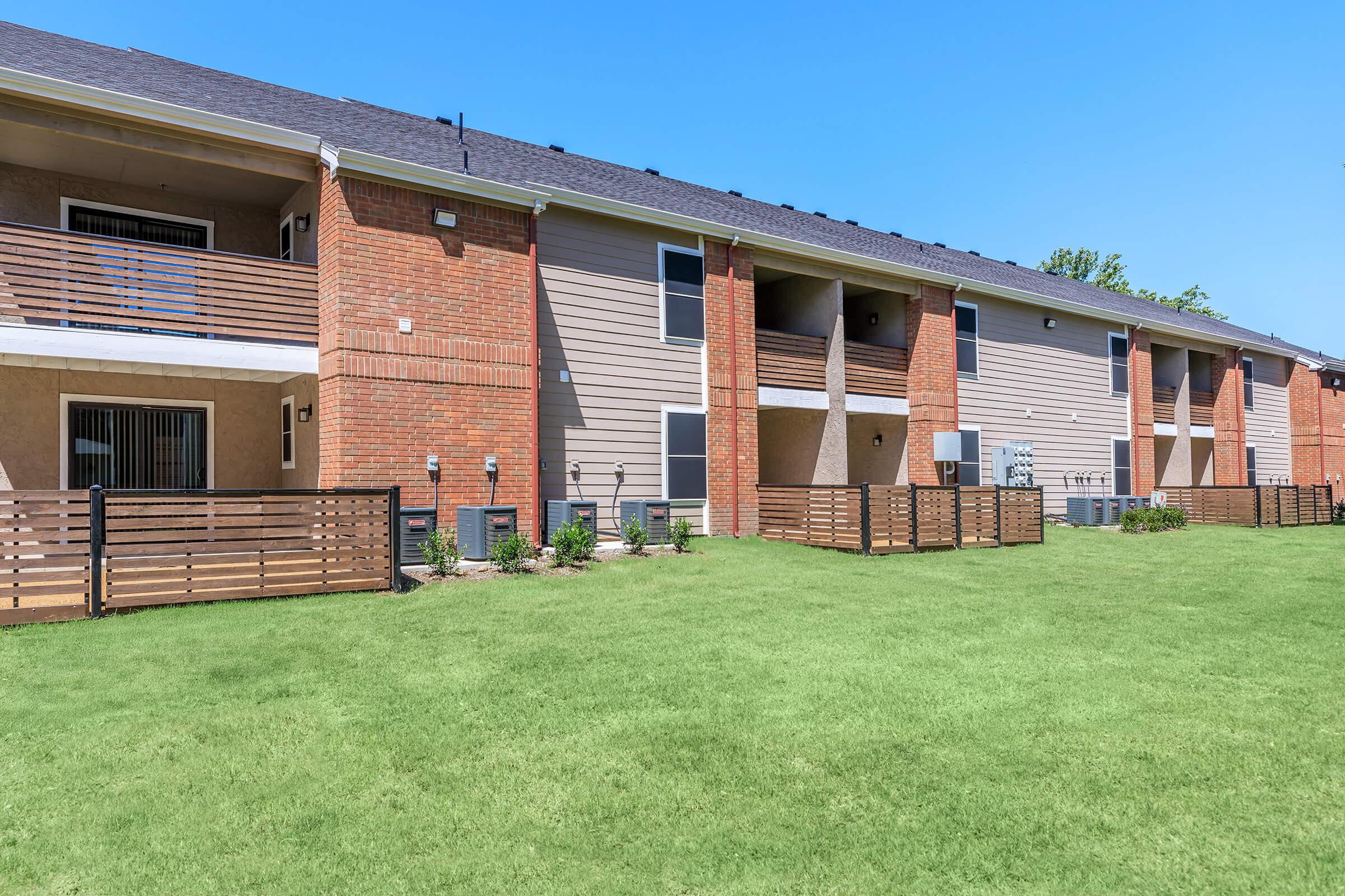 Two-story apartment buildings with wooden balconies and a well-maintained grassy area in front. The buildings feature a mix of brick and siding, with air conditioning units visible. Clear blue sky overhead enhances the bright, inviting atmosphere of the residential complex.
