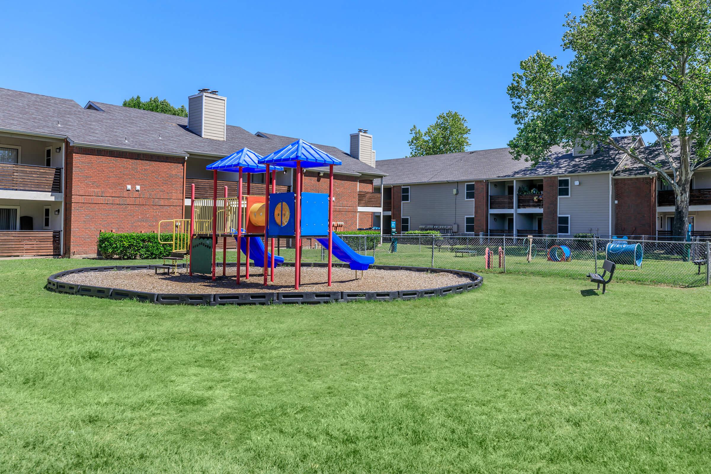 Colorful playground structure with slides and climbing features surrounded by a grassy area. In the background, there are two apartment buildings, one with a brick facade and the other with a light-colored exterior. Clear blue sky overhead, creating a bright outdoor scene.