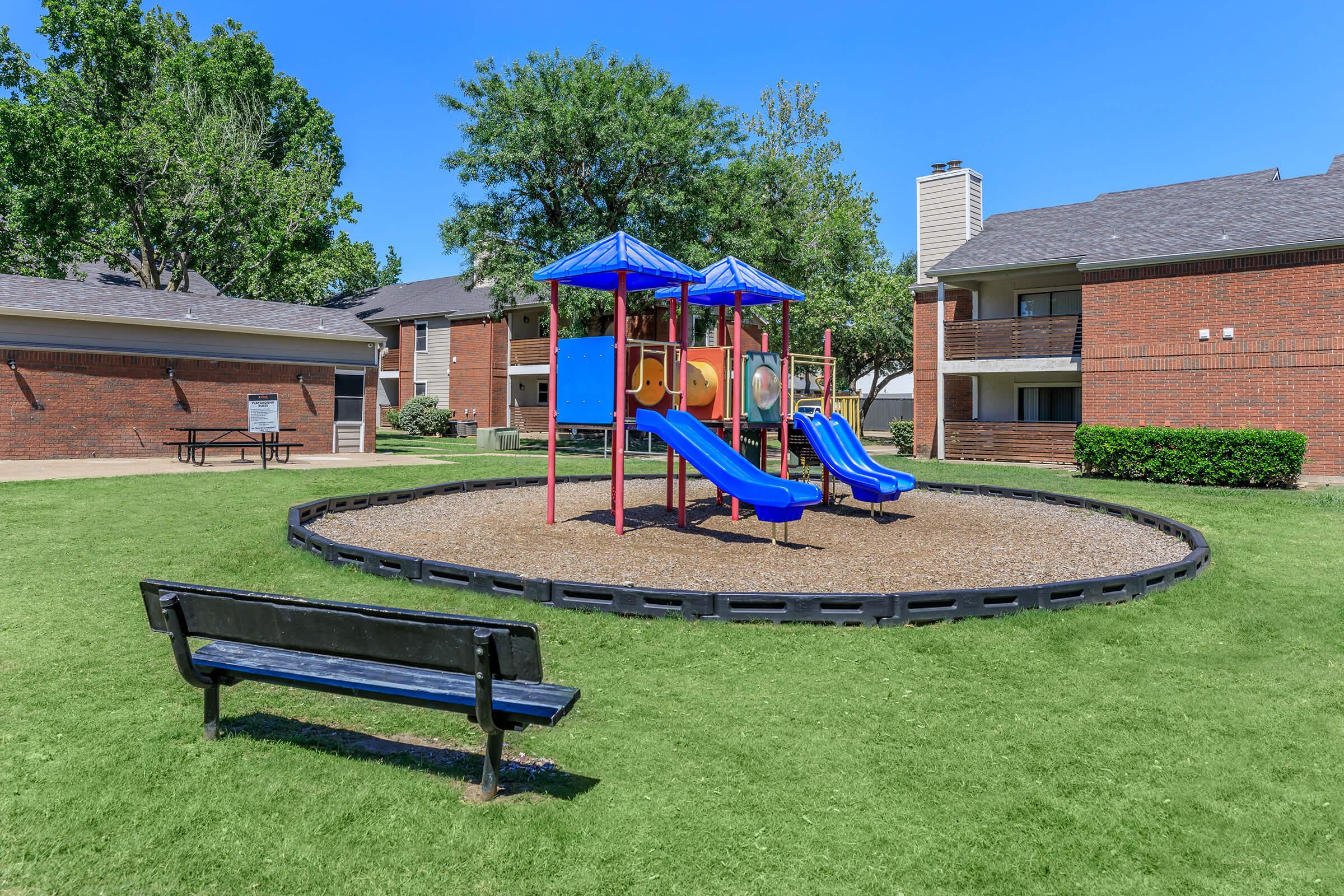 A colorful playground featuring two blue slides and various climbing structures is situated on a grassy area. A black bench is nearby, and the background includes residential apartment buildings and trees under a clear blue sky.