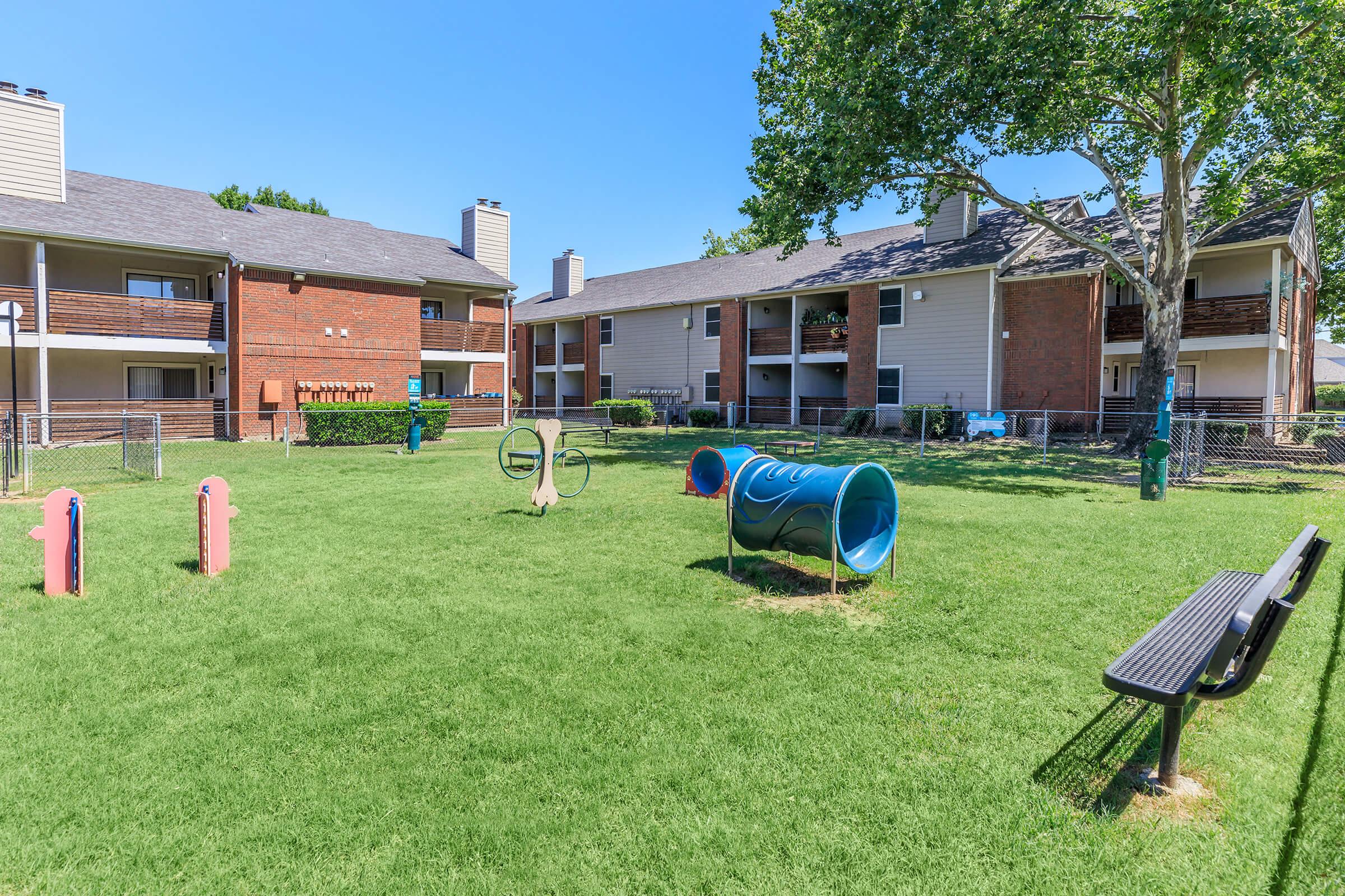 A playground area with bright colored equipment, including tunnels and hoops, surrounded by green grass. In the background, there are two apartment buildings with balconies. The scene is sunny and inviting, with a bench nearby for visitors to sit.