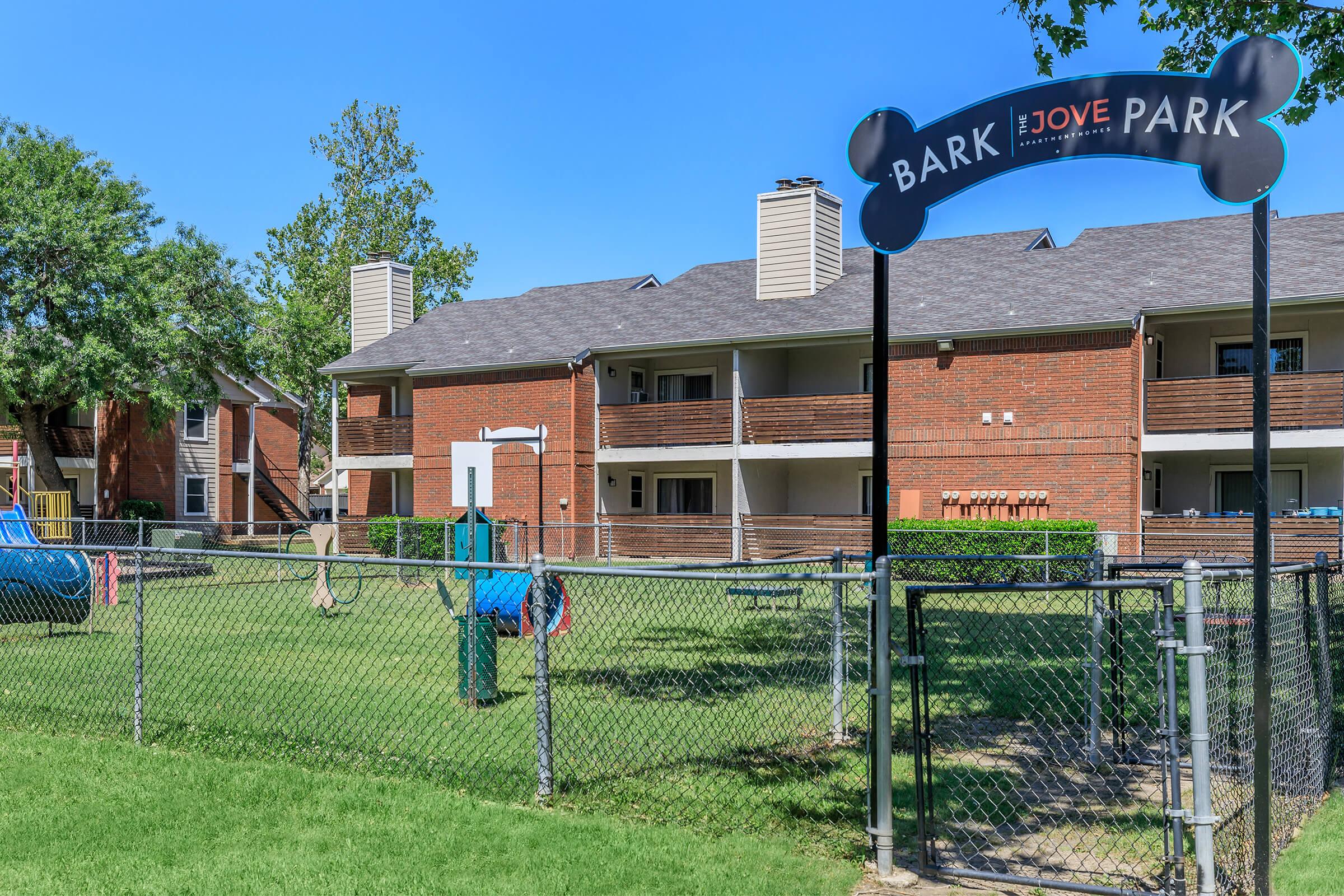 A fenced dog park with a sign that reads "Bark Park" at the entrance, surrounded by grassy areas. The background features a two-story brick apartment building with balconies and trees providing shade. There are play structures and dog-friendly amenities visible within the park.