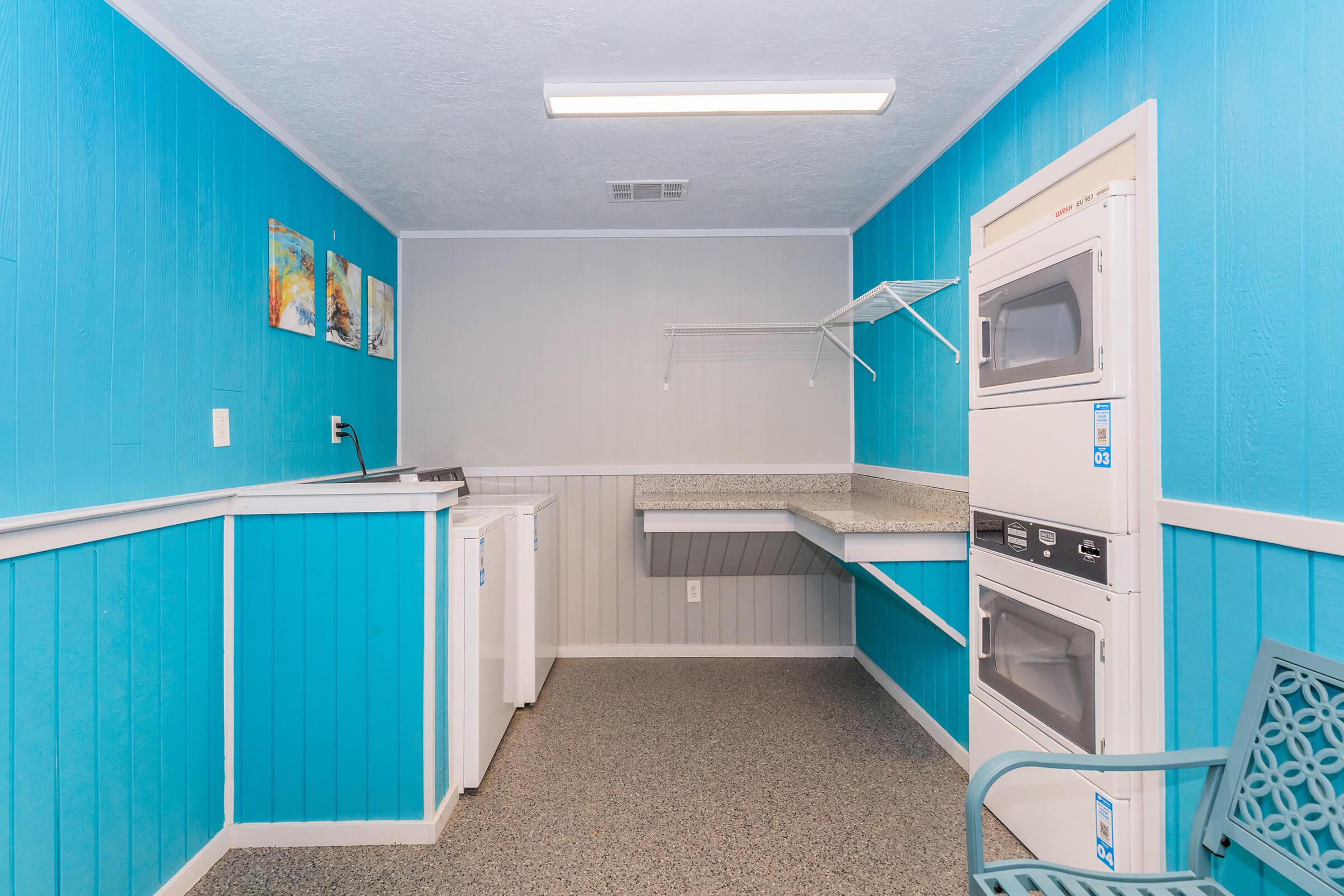 A bright laundry room with blue walls and gray accents. It features a washing machine and dryer stacked on one side, a countertop for folding, and a small seating area with a teal chair. There are decorative paintings on the wall and shelves for storage above the countertop.