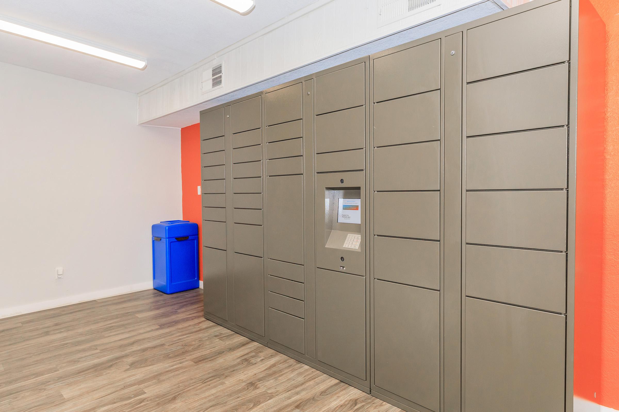 A row of metallic package lockers against an orange wall in a well-lit room. A keypad and screen are visible on one of the lockers, and a blue trash bin is placed nearby. The flooring features light wood-like panels, creating a modern and functional space for package retrieval.