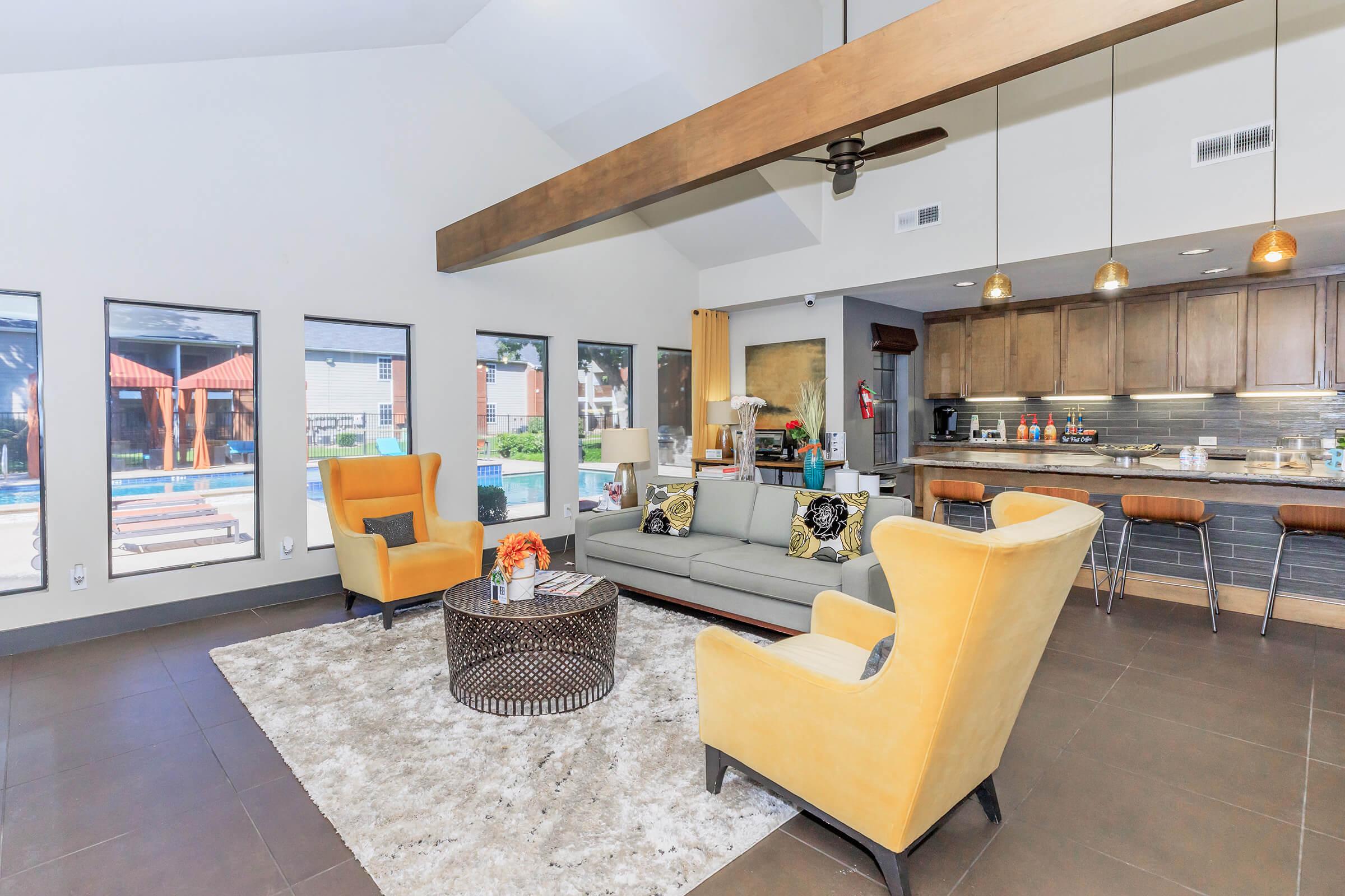 Modern living room with a light gray couch and two yellow armchairs. A round coffee table sits on a plush rug in the center. Large windows offer views of an outdoor area, and a kitchen with wooden cabinetry and bar seating is visible in the background. The space is bright and inviting.