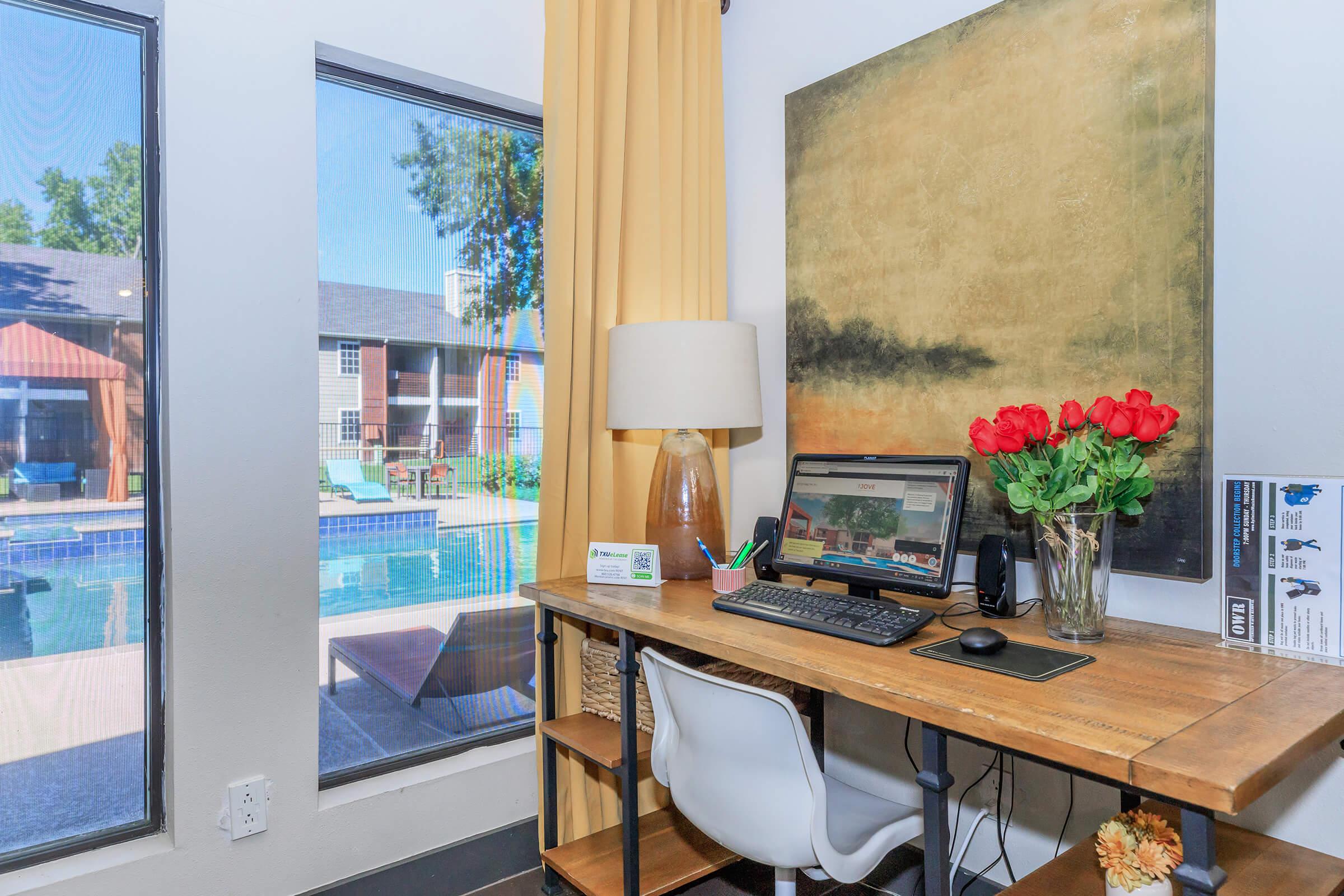 A cozy workspace featuring a wooden desk with a computer, a lamp, and a vase of red roses. Large windows provide a view of a pool area outside, surrounded by greenery and lounge chairs. Warm yellow curtains add a touch of color to the room.