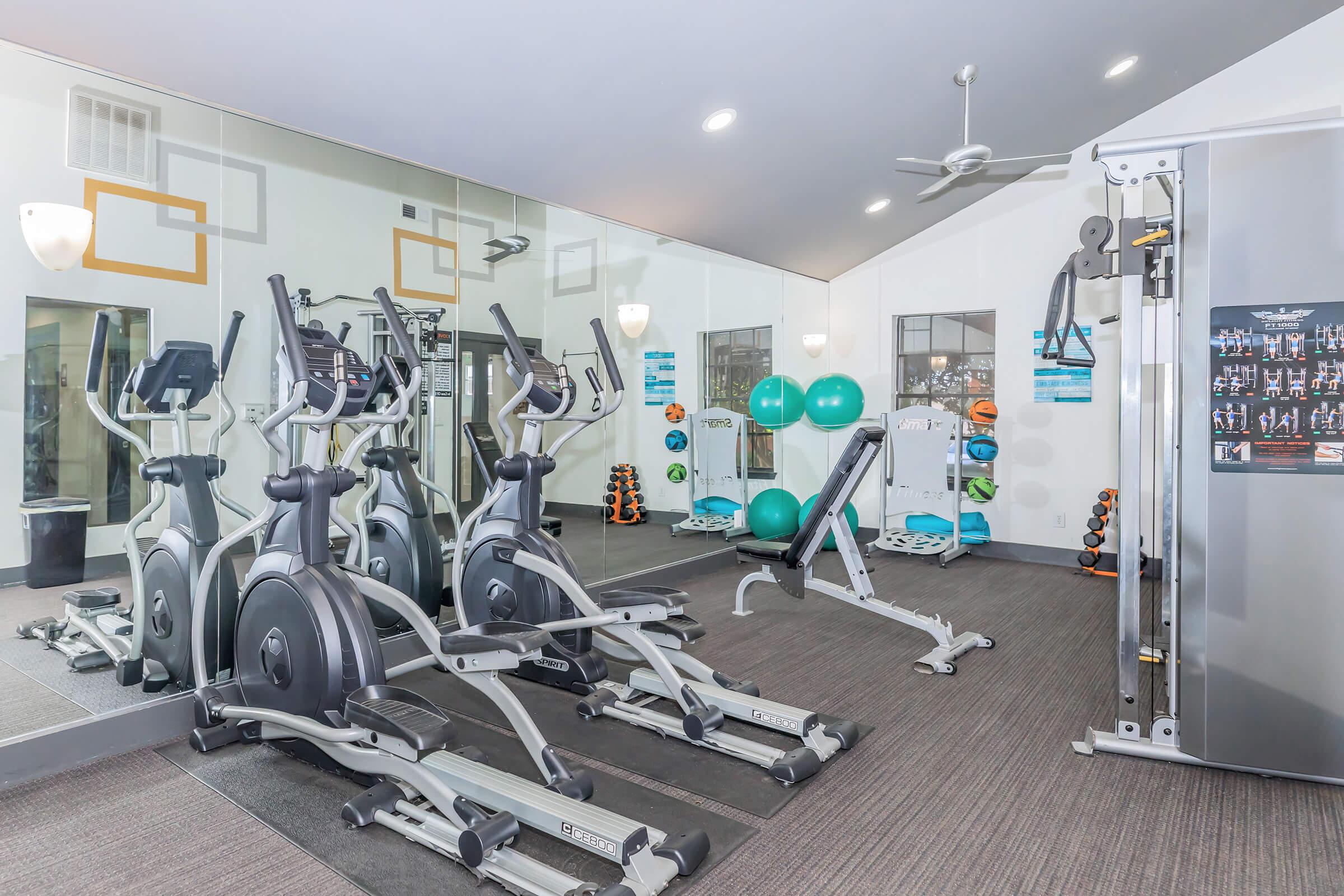 A modern gym interior featuring several pieces of exercise equipment, including elliptical machines, a weight bench, and a multi-gym machine. Bright lights and large mirrors enhance the spacious feel, while exercise balls and weights are neatly arranged in the background. The environment promotes fitness and wellness.