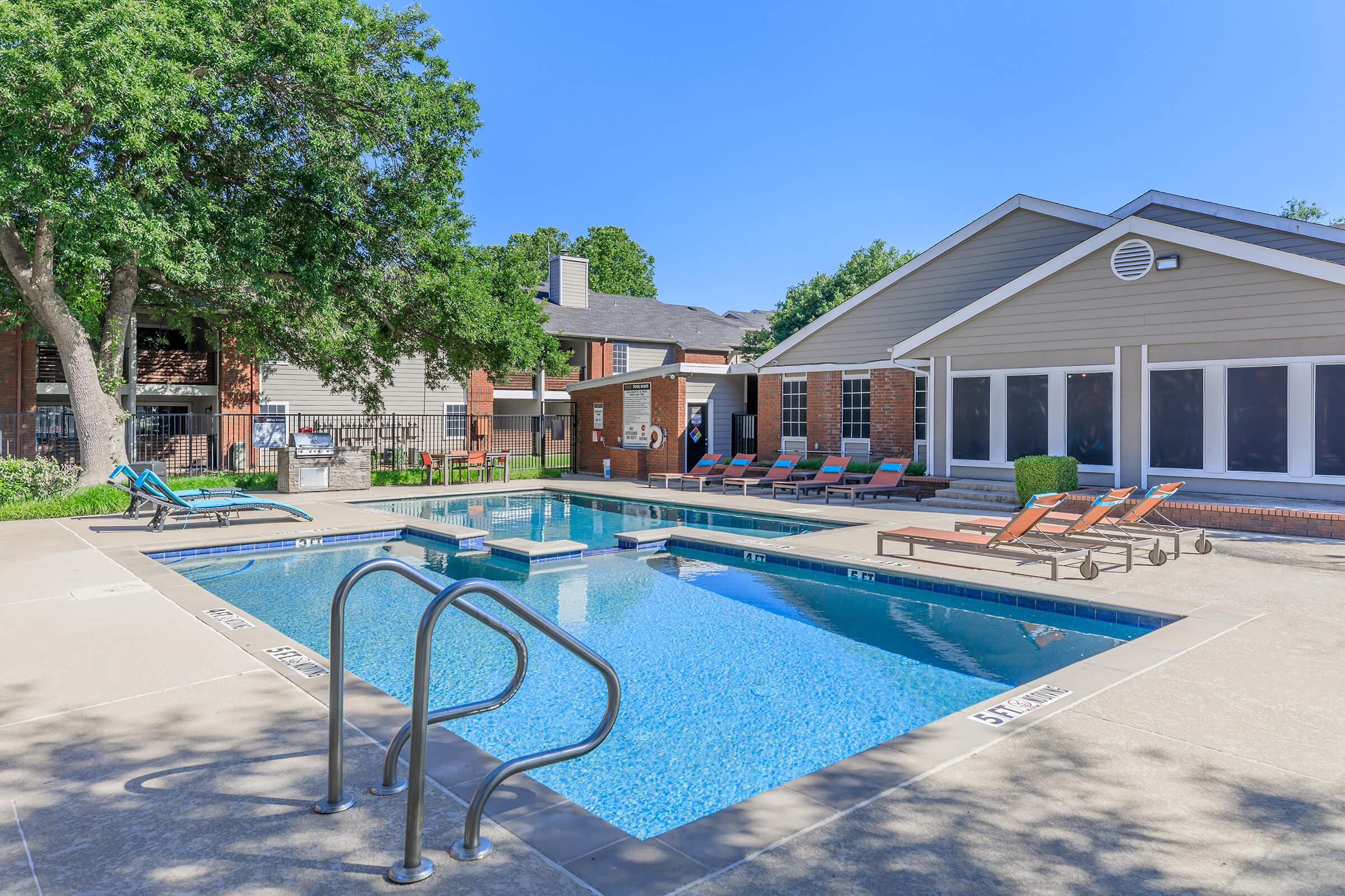 A bright outdoor swimming pool surrounded by lounge chairs and shaded by trees. Nearby, there are residential buildings with large windows. The scene is clear and sunny, showcasing a relaxing atmosphere ideal for leisure activities.