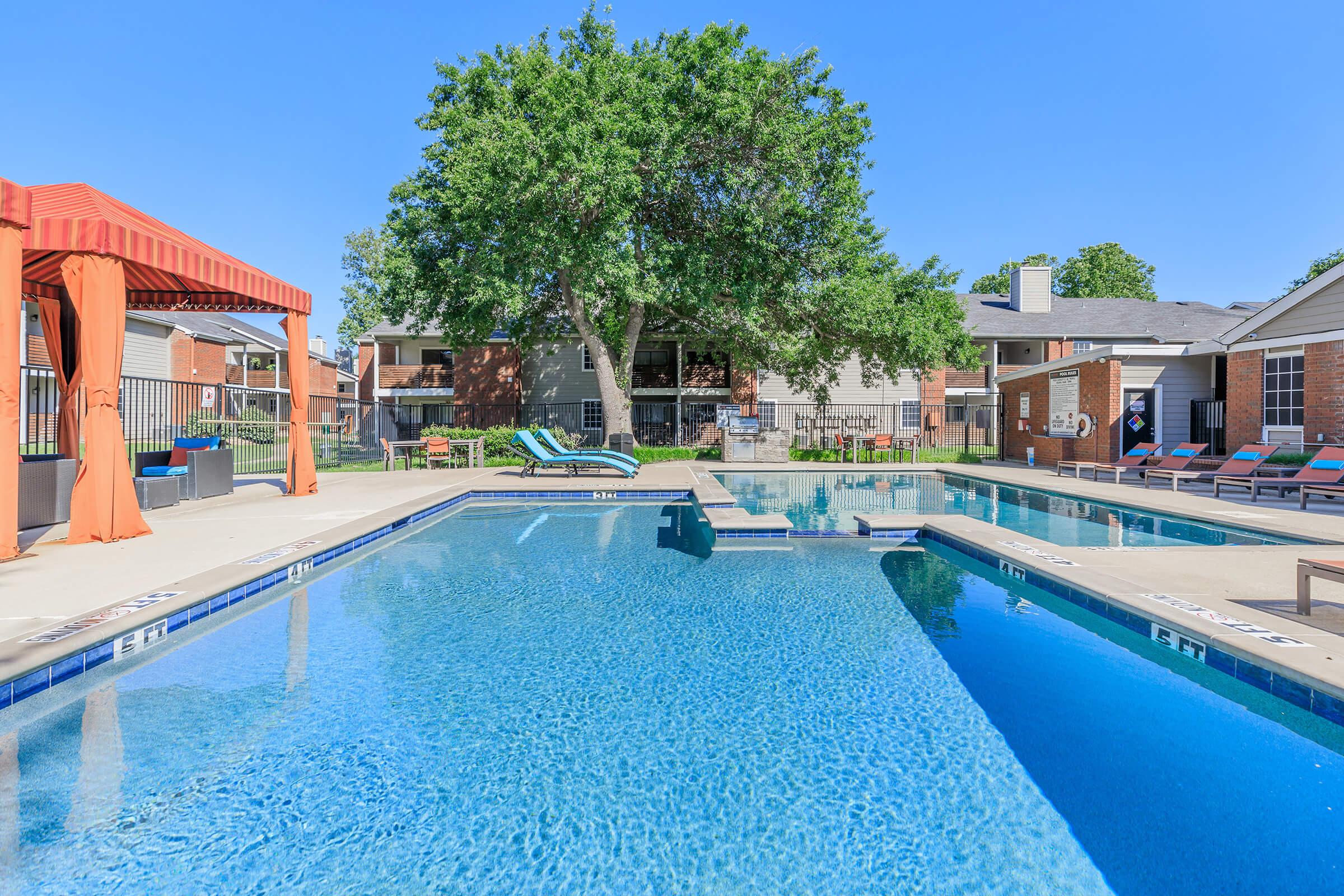 A clear, sunny day at a well-maintained pool area featuring two swimming pools, lounge chairs, and a shaded cabana. Surrounding the pools are trees and residential buildings, creating a relaxing atmosphere for leisure and social activities.