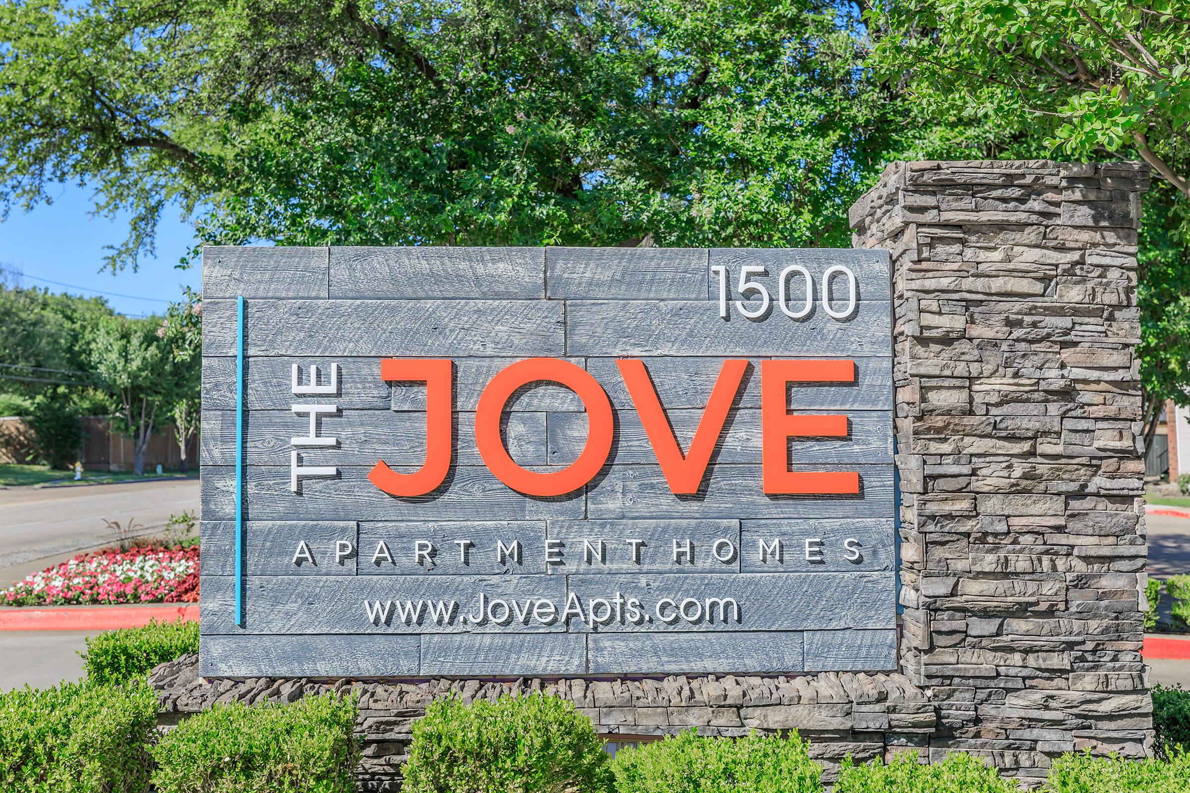 Signage for "The Jove Apartment Homes" displaying the address "1500." The sign features a modern design with orange and gray colors, surrounded by greenery and shrubs. The website is also included: www.JoveApts.com.