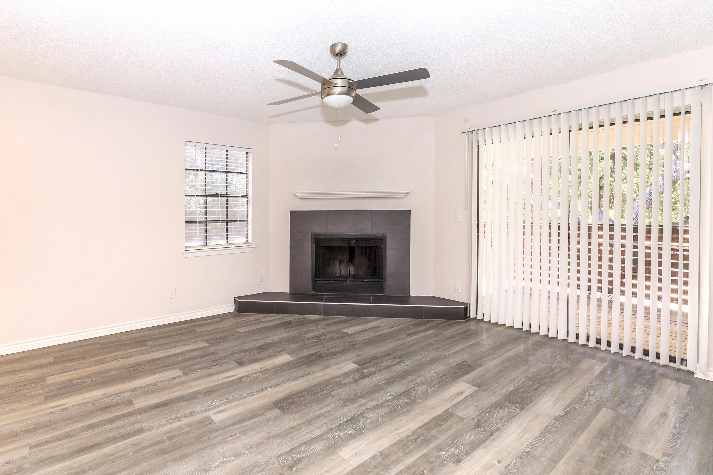 A spacious living room featuring a ceiling fan, a black fireplace set into the wall, and large windows with vertical blinds. The flooring is a light wood laminate, creating a bright and airy atmosphere, with neutral wall colors enhancing the openness of the space.