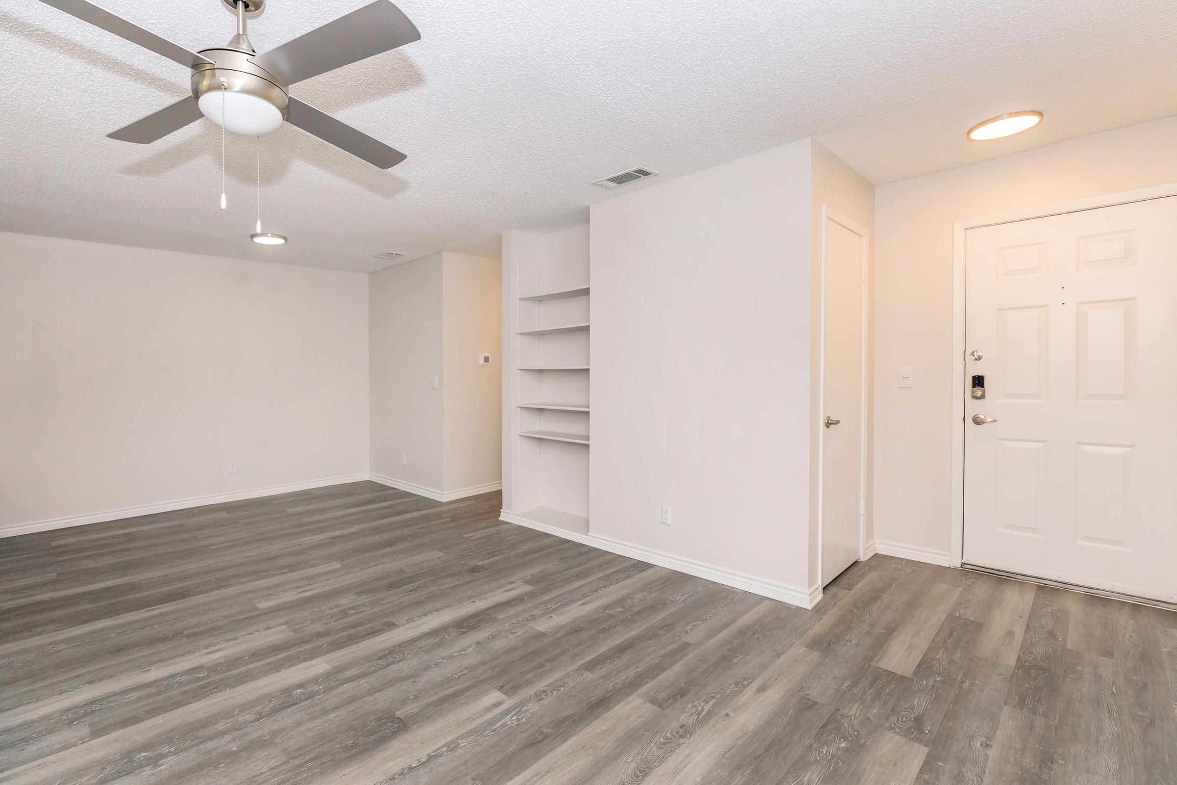 A spacious living room area featuring light-colored walls, a ceiling fan, laminate flooring, and a built-in shelf. A door on the right leads outside, and there is ample natural light coming in. The space is clean and empty, suitable for personalized decoration.