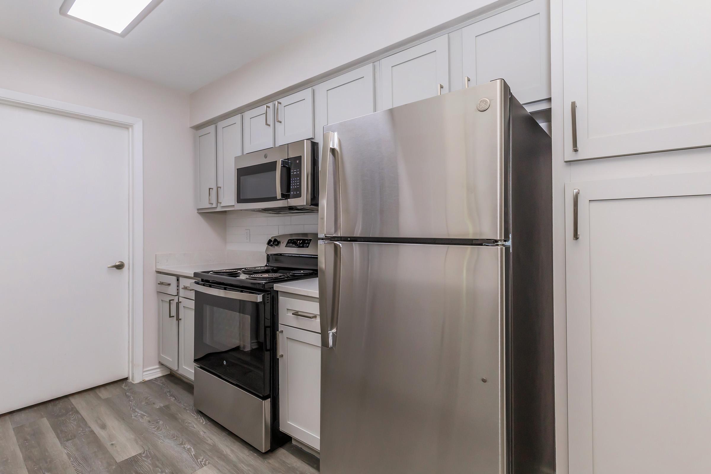 Interior view of a modern kitchen featuring stainless steel appliances, including a refrigerator, microwave, and oven. The cabinetry is light gray, and the countertop is white, complemented by light wood-like flooring. A door in the background leads to another room.