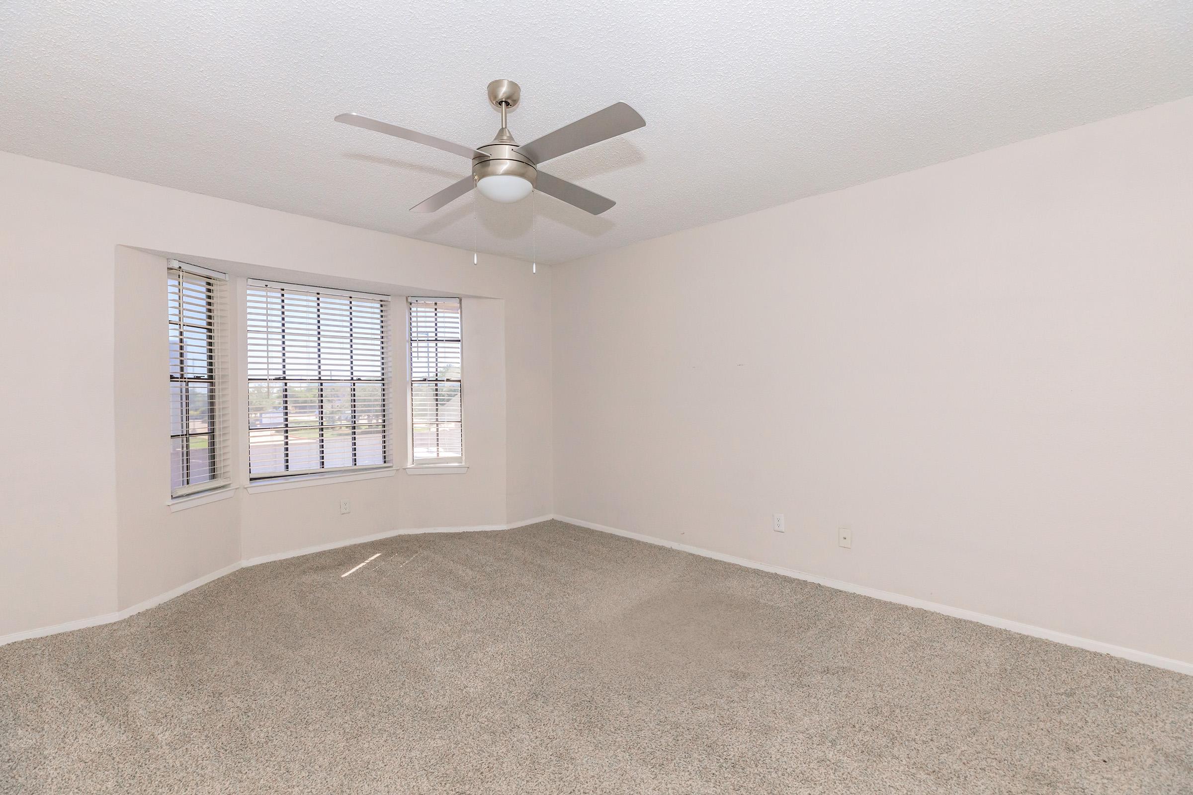 A spacious empty room with light beige walls and new carpet. It features a ceiling fan and a large bay window with blinds, allowing natural light to enter. The floor is clean and the overall atmosphere is bright and inviting. Ideal for customization and decoration.