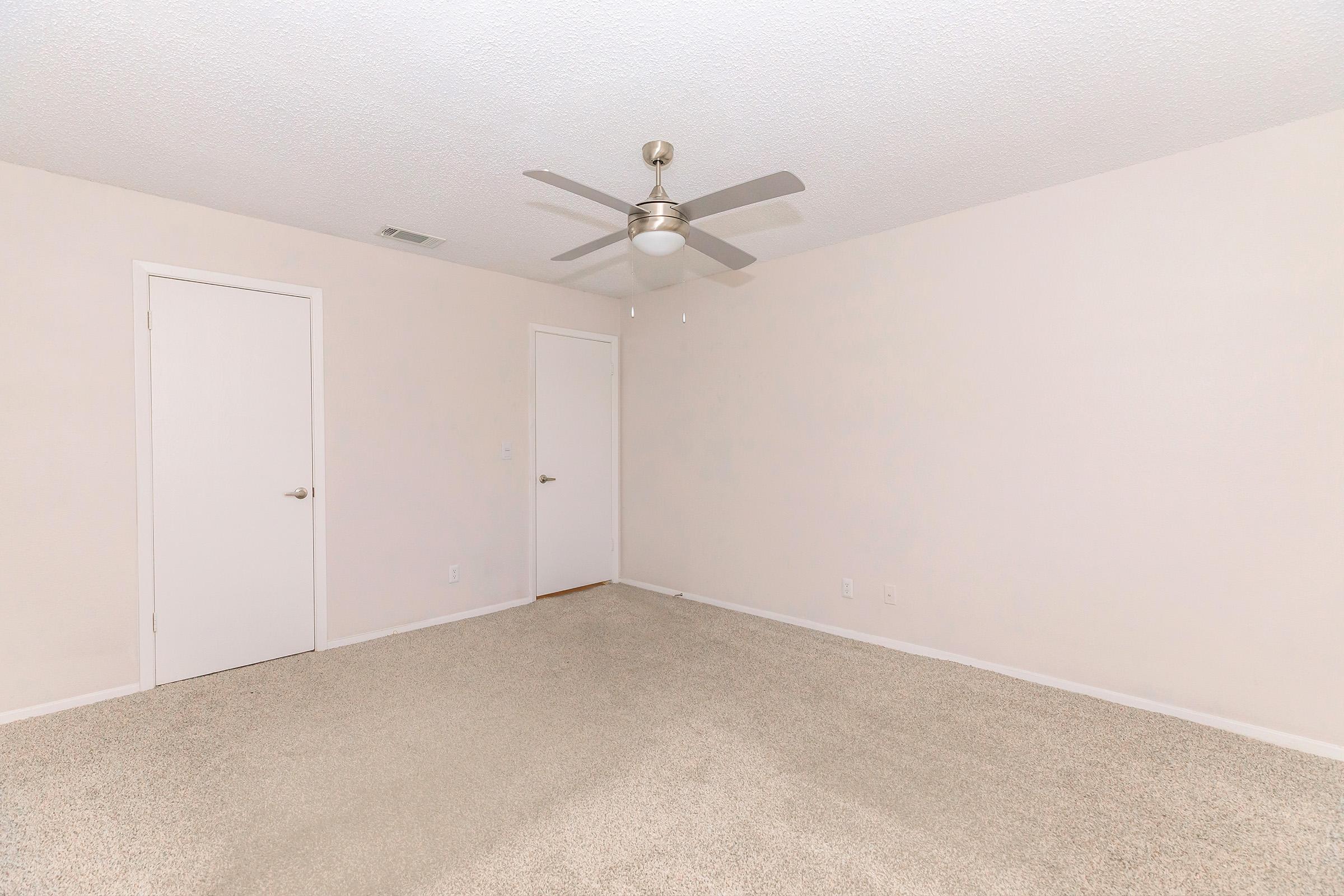 A spacious, empty room with light beige carpet flooring. There are two closed white doors on opposite walls and a ceiling fan in the center. The walls are painted a soft off-white color, contributing to a bright and airy atmosphere.