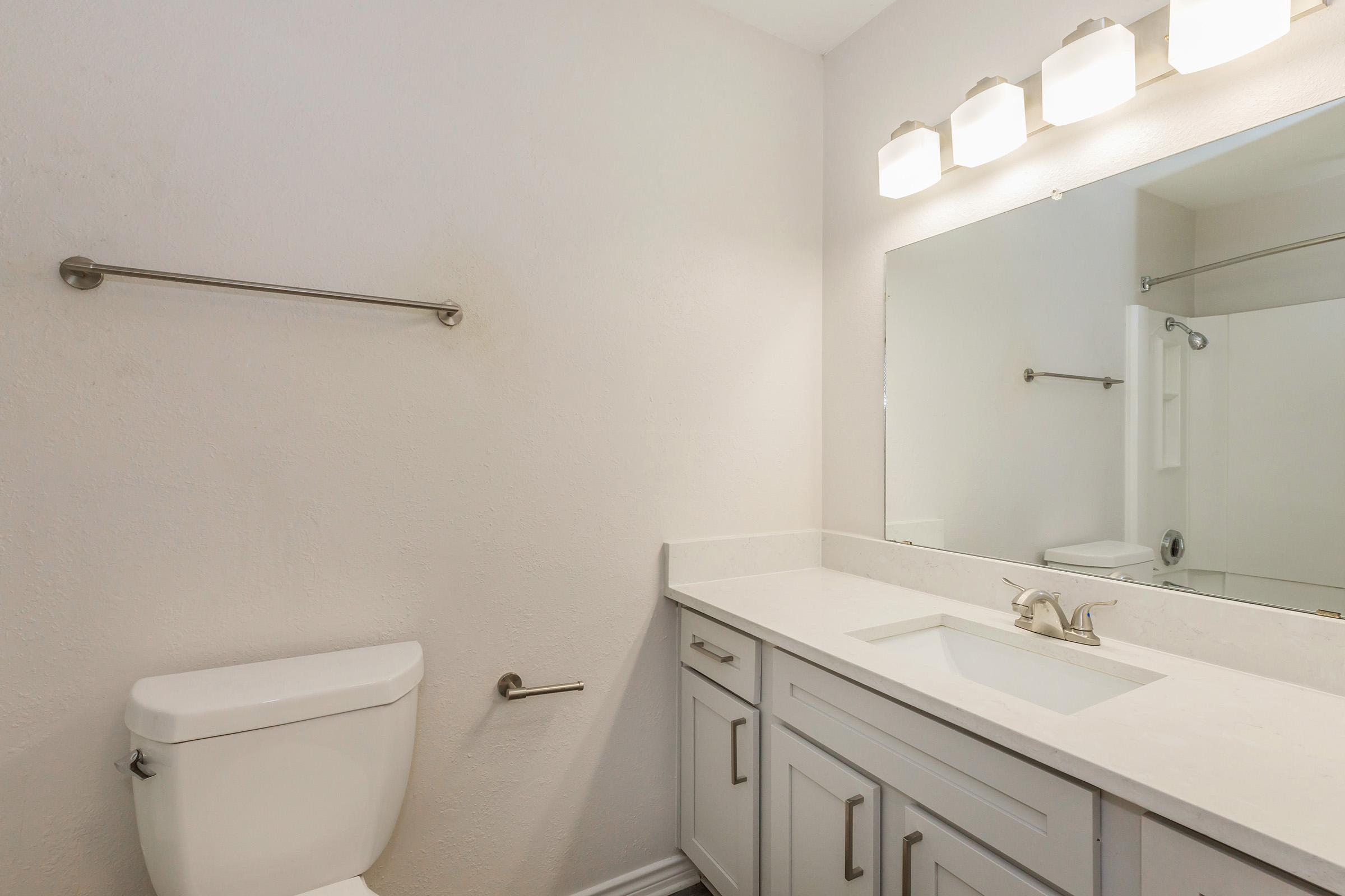 A modern bathroom featuring a white countertop with a sink, a large mirror above, and a light fixture with three bulbs. On the left, there is a white toilet, and a towel bar is mounted on the wall. A shower area is visible in the corner, with a towel hanging nearby. Walls are light-colored, creating a clean and spacious feel.