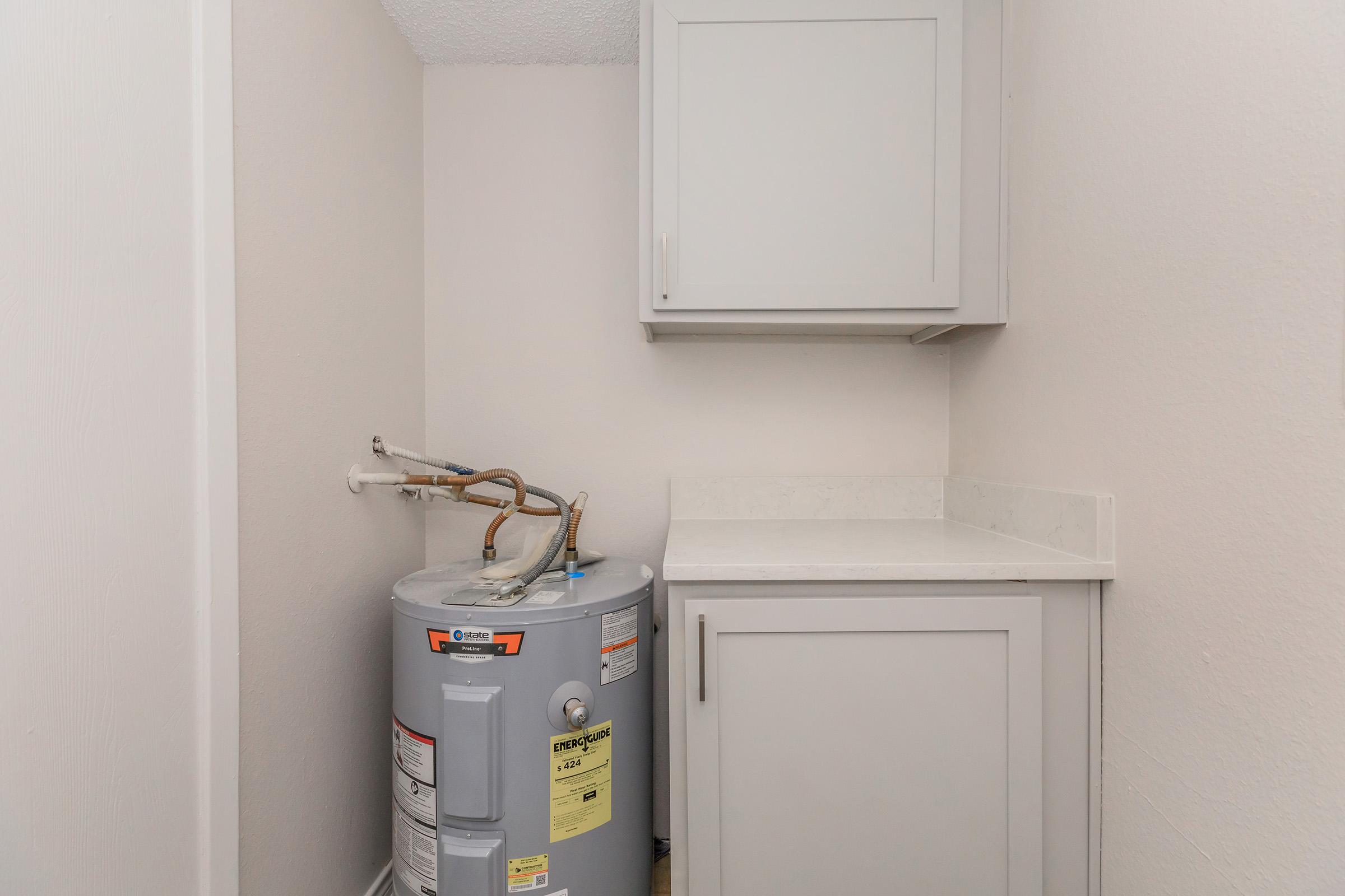 A small laundry space featuring a water heater with visible pipes and a cabinet above. Adjacent to the water heater is a countertop, all set against a light-colored wall. The area is neatly organized, with no clutter visible.