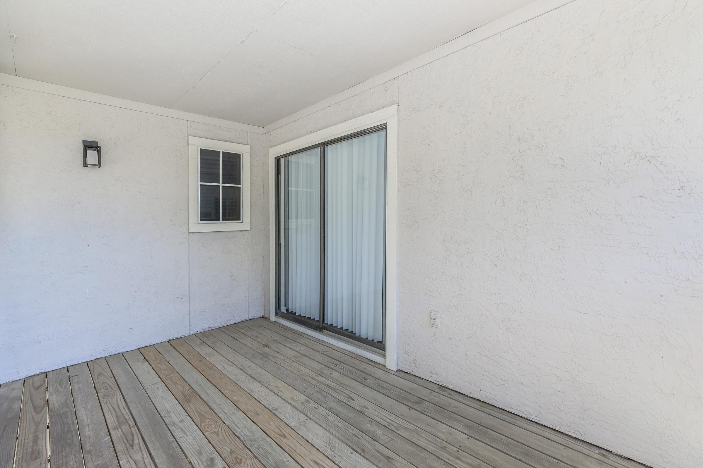 A simple balcony or patio area featuring a wooden floor, a white stucco wall, a sliding glass door with vertical blinds, and a small window with a grid pattern. The space is well-lit and has minimal furnishings, offering an open and clean atmosphere.