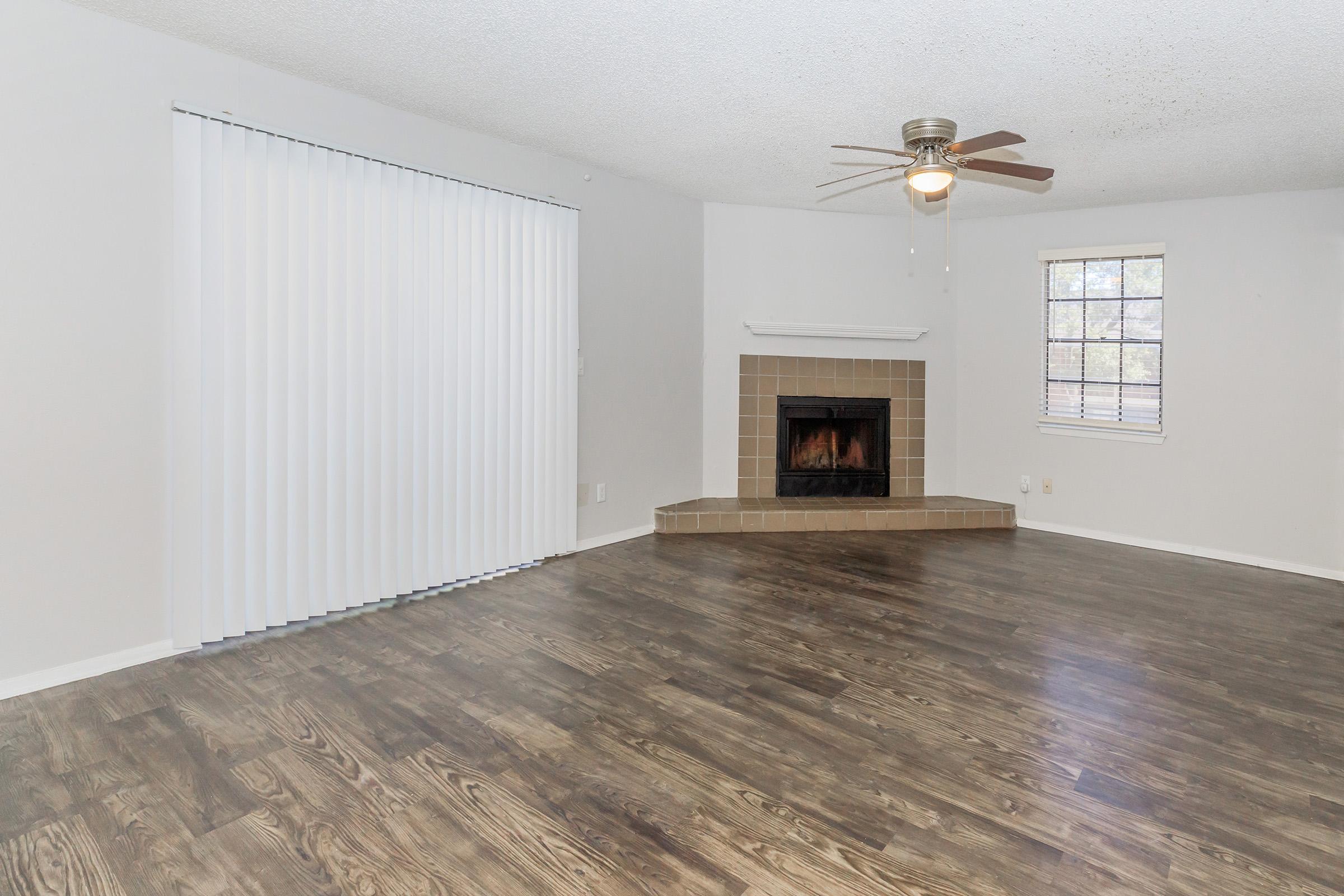 Spacious living room featuring a fireplace with a tiled surround, wooden flooring, and large windows with vertical blinds. The walls are painted in a light color, creating an airy atmosphere. A ceiling fan adds to the comfort of the space.